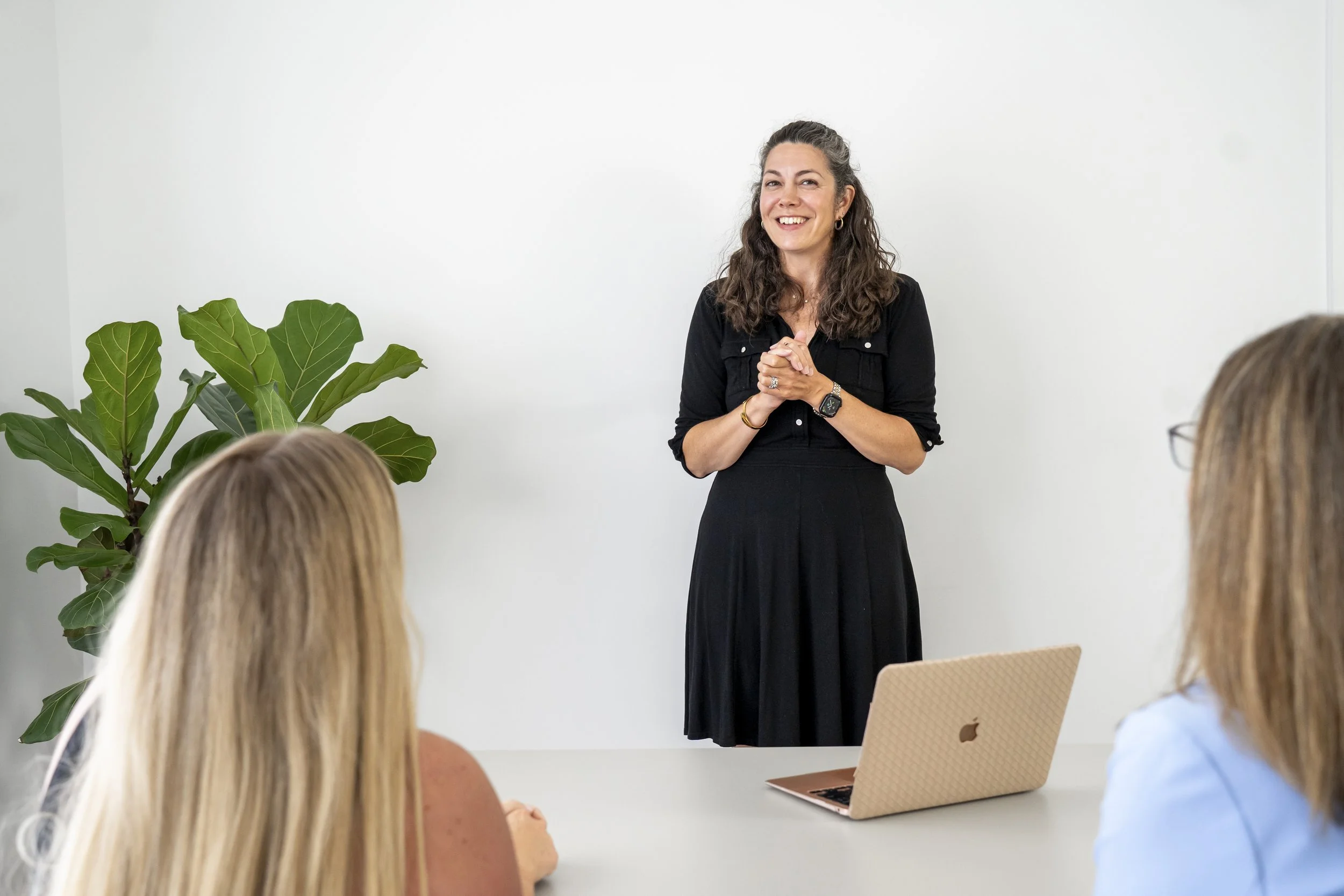 Katie Chew in a black dress presenting to colleagues in a modern office, with a laptop and large houseplant nearby