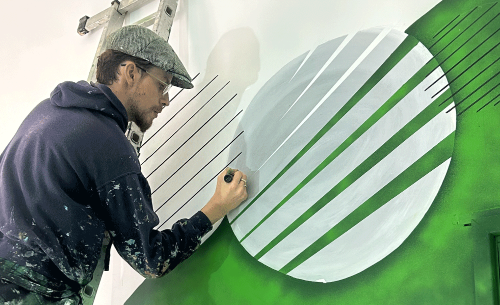A man wearing glasses, a gray flat cap, and a navy hoodie with paint splatters is painting a mural on a white wall. The mural features a large white circle with green and black lines, resembling a stylized planet or abstract design.