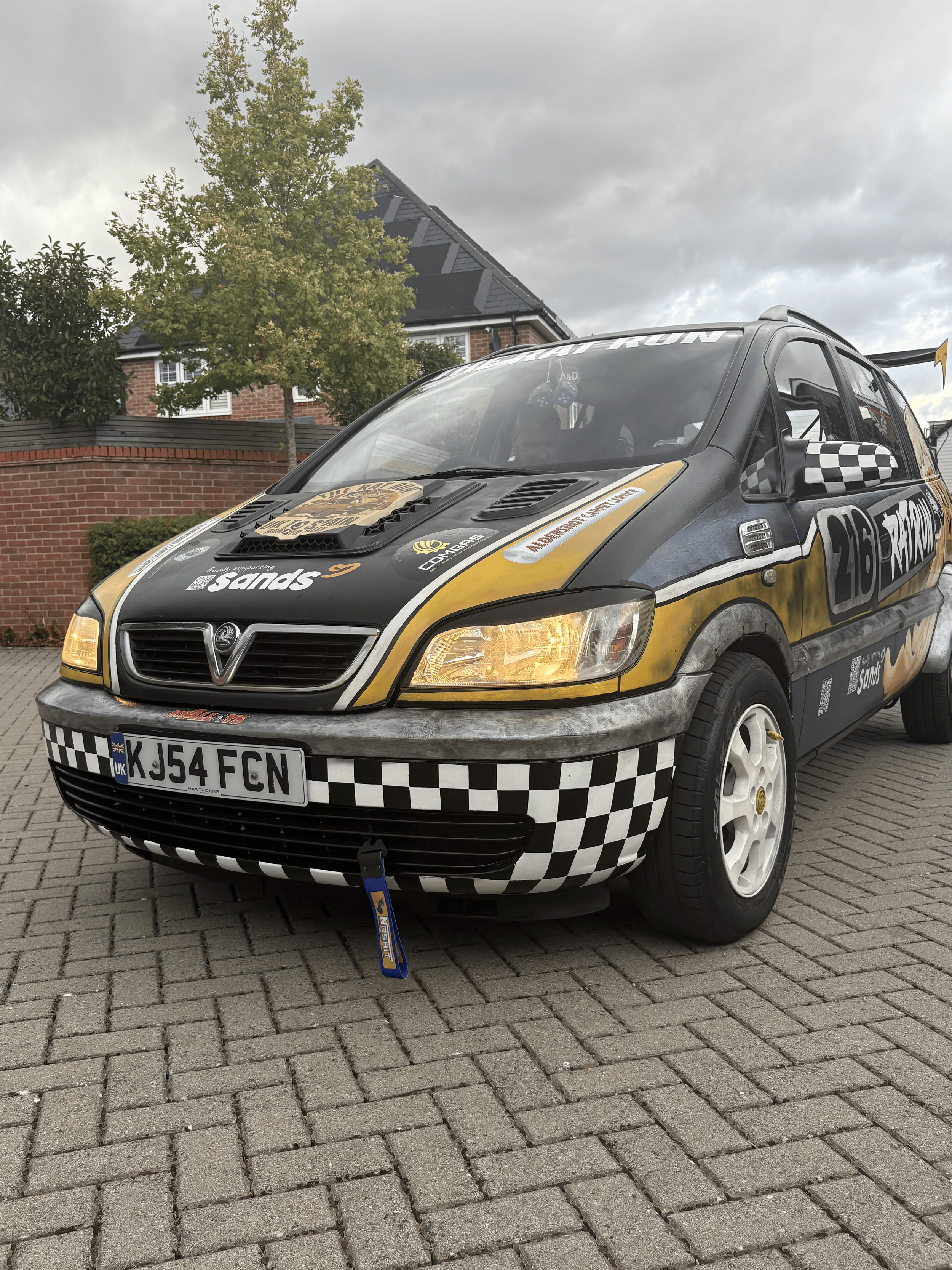 Close-up of a race car with a black, gold, and white checkered design, parked on a cobblestone driveway, with a brick house, a tree, and a cloudy sky in the background.