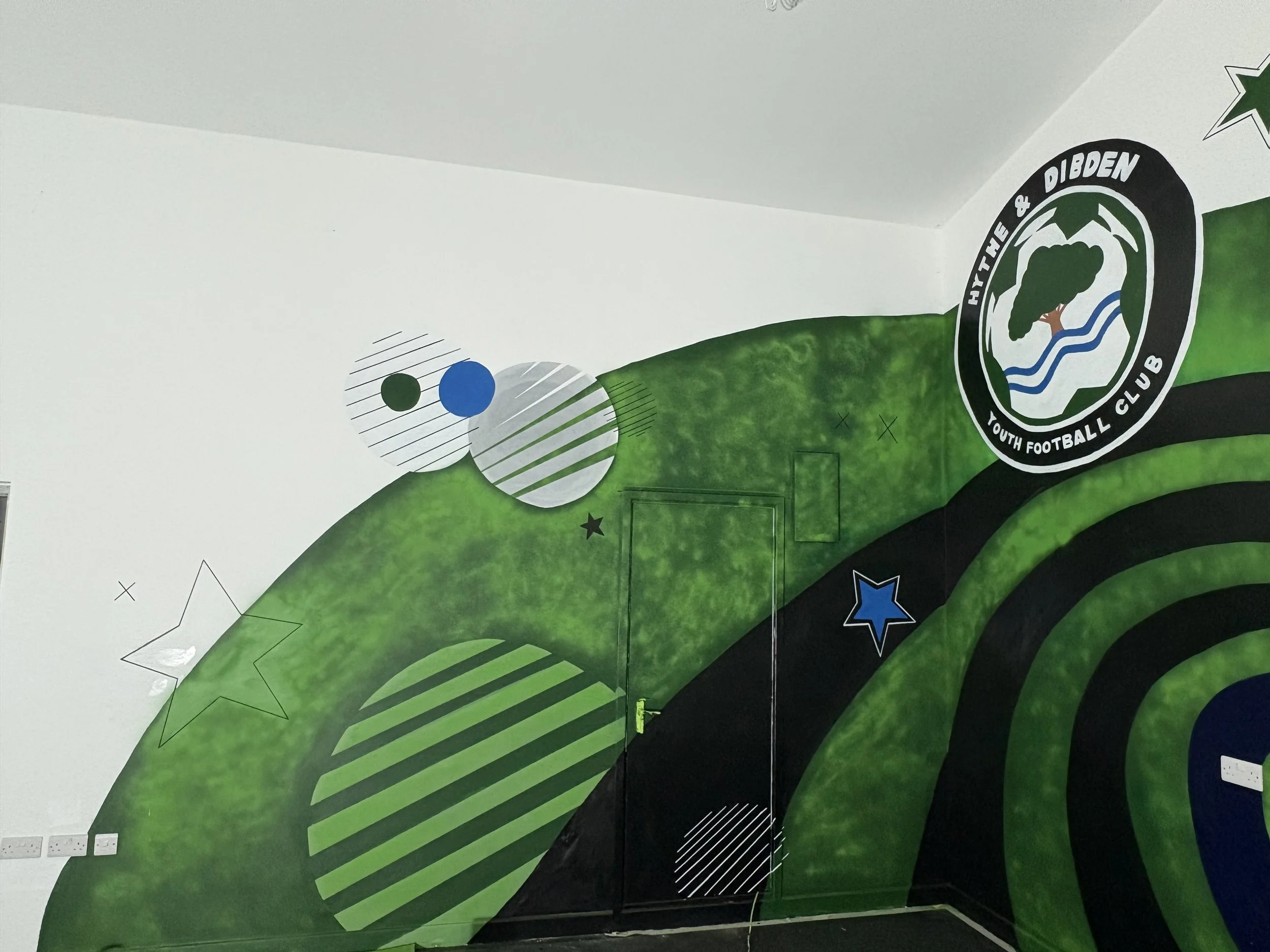 A mural with a football club badge that reads "HYT THE & DIBDEN YOUTH FOOTBALL CLUB". The mural features green, black, and white colors with stars, circles, and stripe designs.