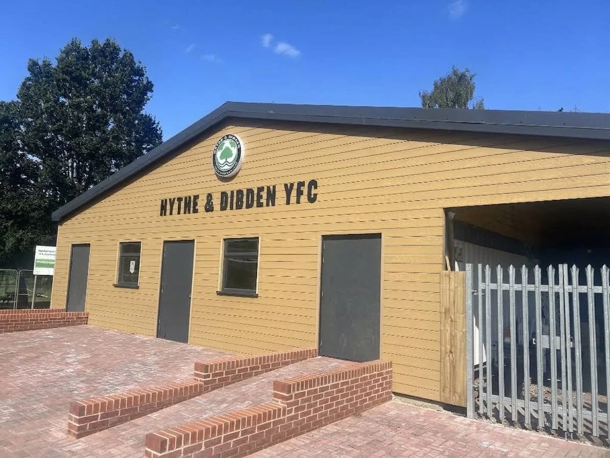 A yellow wooden building with the sign 'HYTHE & DIBDEN YFC' and a round logo at the top. The building has three dark doors and three small windows. There is a brick-paved area in front with a low brick wall. A wooden gate and fencing are on the right side. Trees are visible in the background under a clear blue sky.