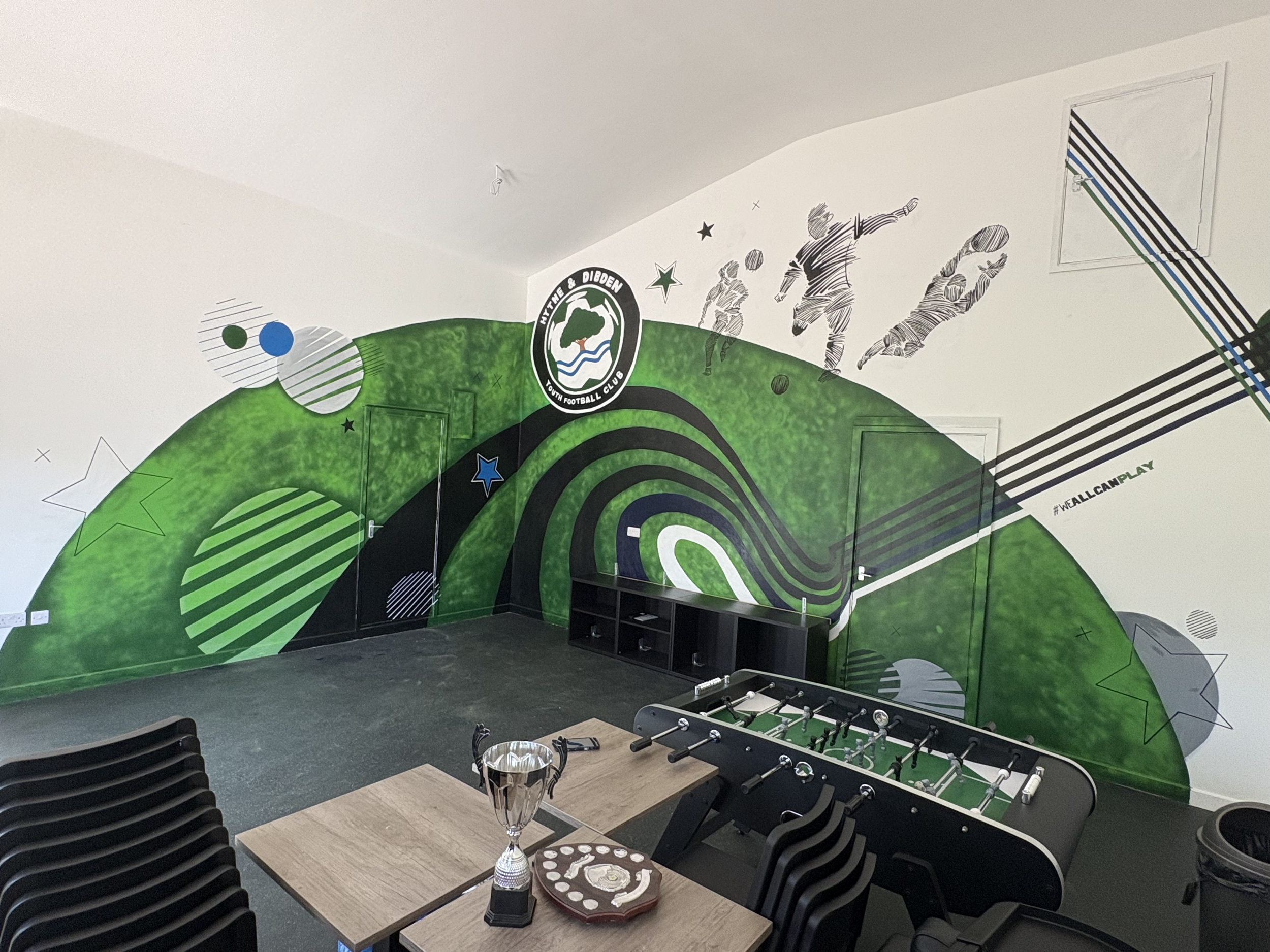 Indoor room with green sports-themed wall mural, featuring soccer players, stars, and the logo of Hyde & Diden Youth Football Club. There is a foosball table, a wooden table with a trophy, a circular plaque, and some chairs.