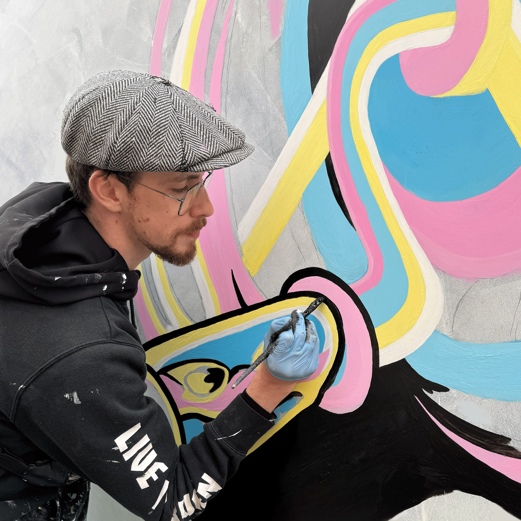 A man wearing glasses, a gray plaid cap, and a black hoodie painting a colorful mural on a wall. The mural features abstract shapes and bright colors including pink, yellow, blue, black, and white.