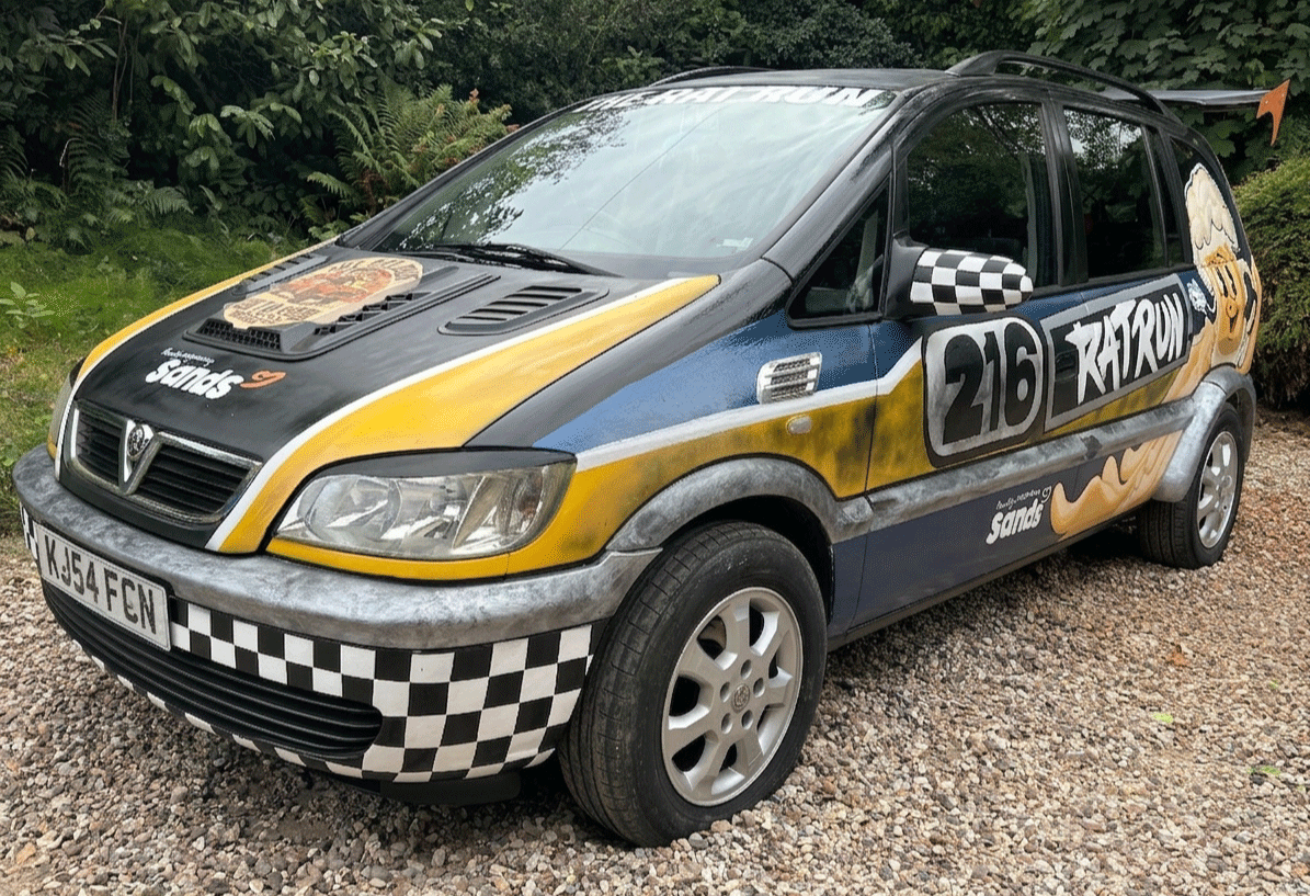 A decorated car with racing-themed graphics and a checkered pattern on the front and side, parked on a gravel surface with greenery in the background.