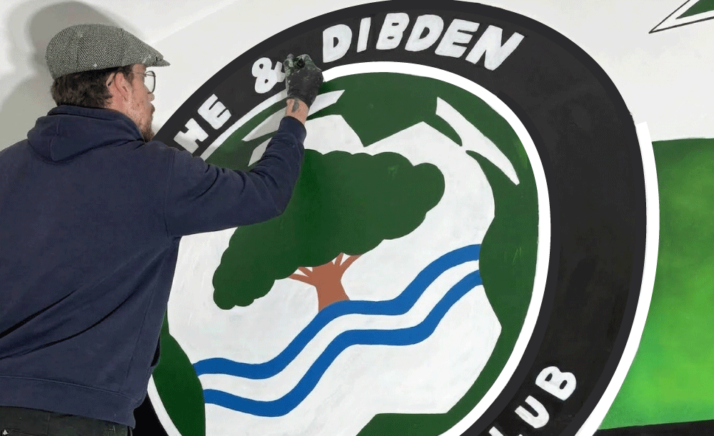 A man painting a circular emblem that features a tree, water, and mountain landscape, with the text "The DIBDEN" around the top.