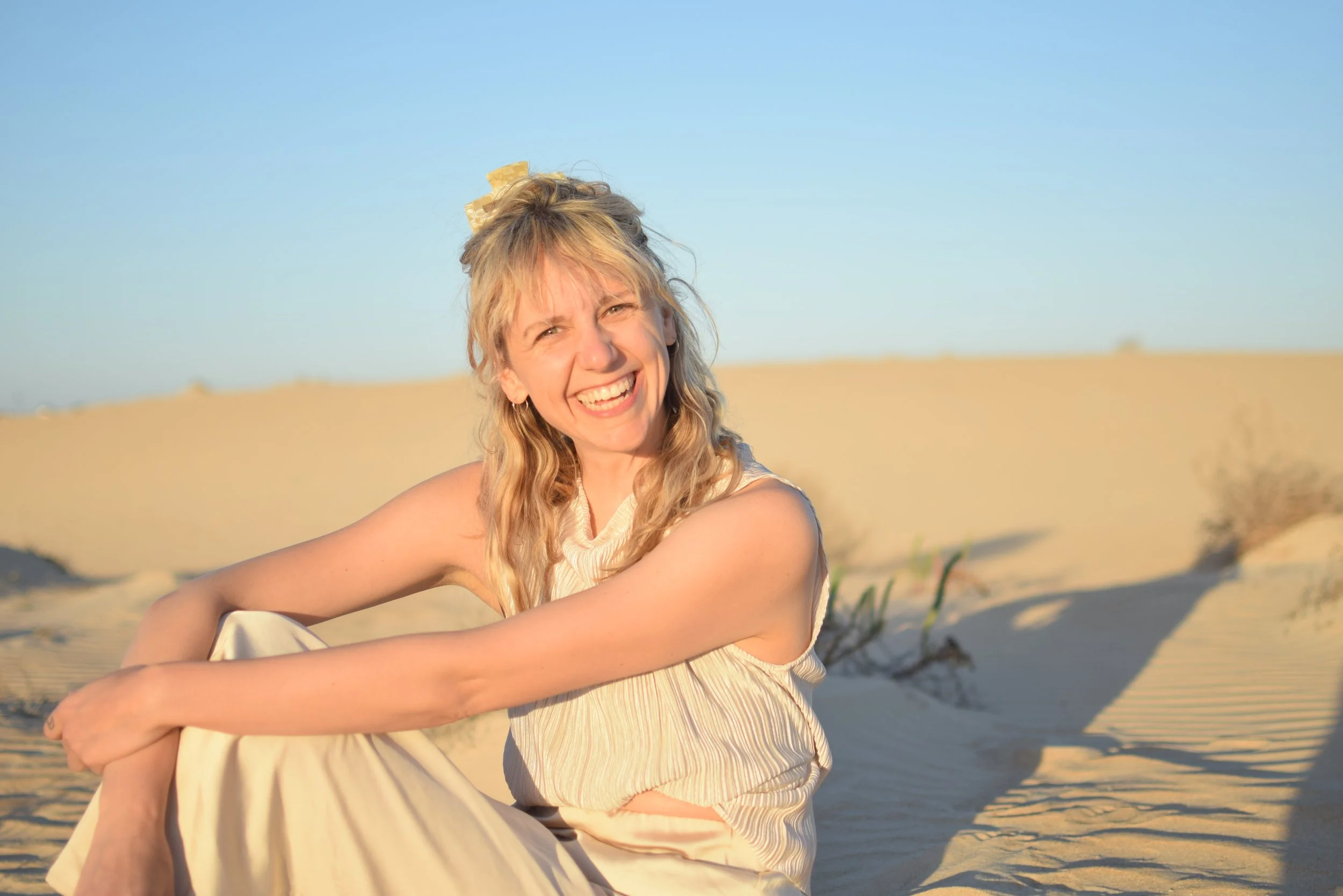A woman with blonde hair sitting and smiling in a desert landscape under a clear blue sky.