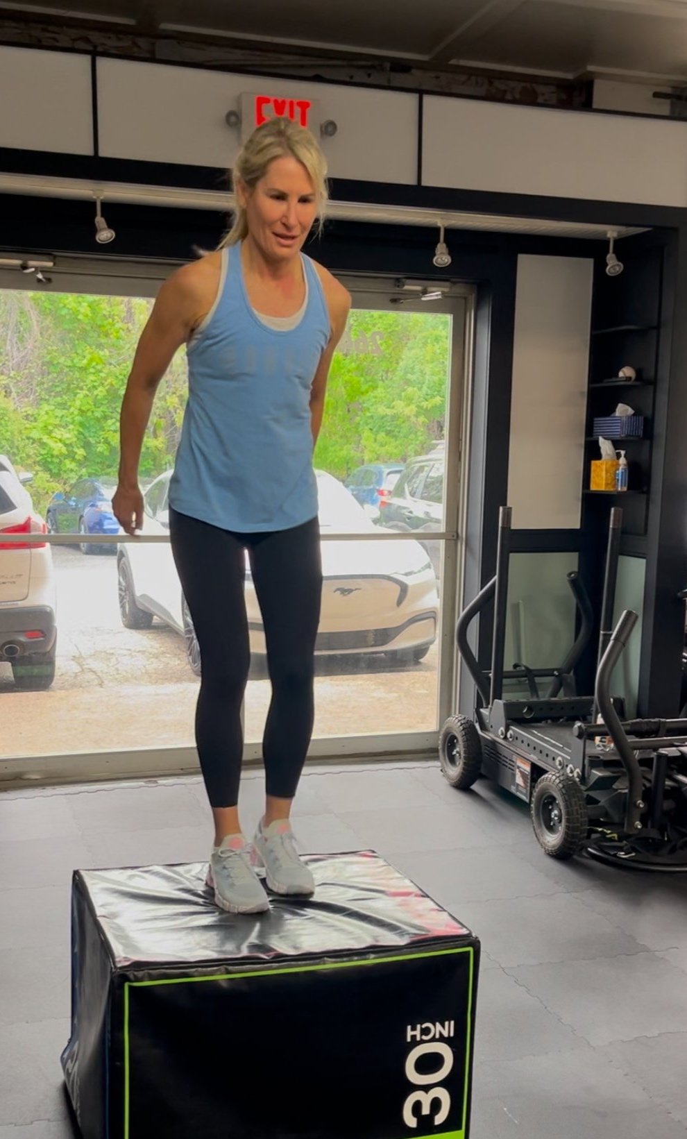 A woman in athletic clothing performing a box jump exercise on a black exercise box inside a gym.