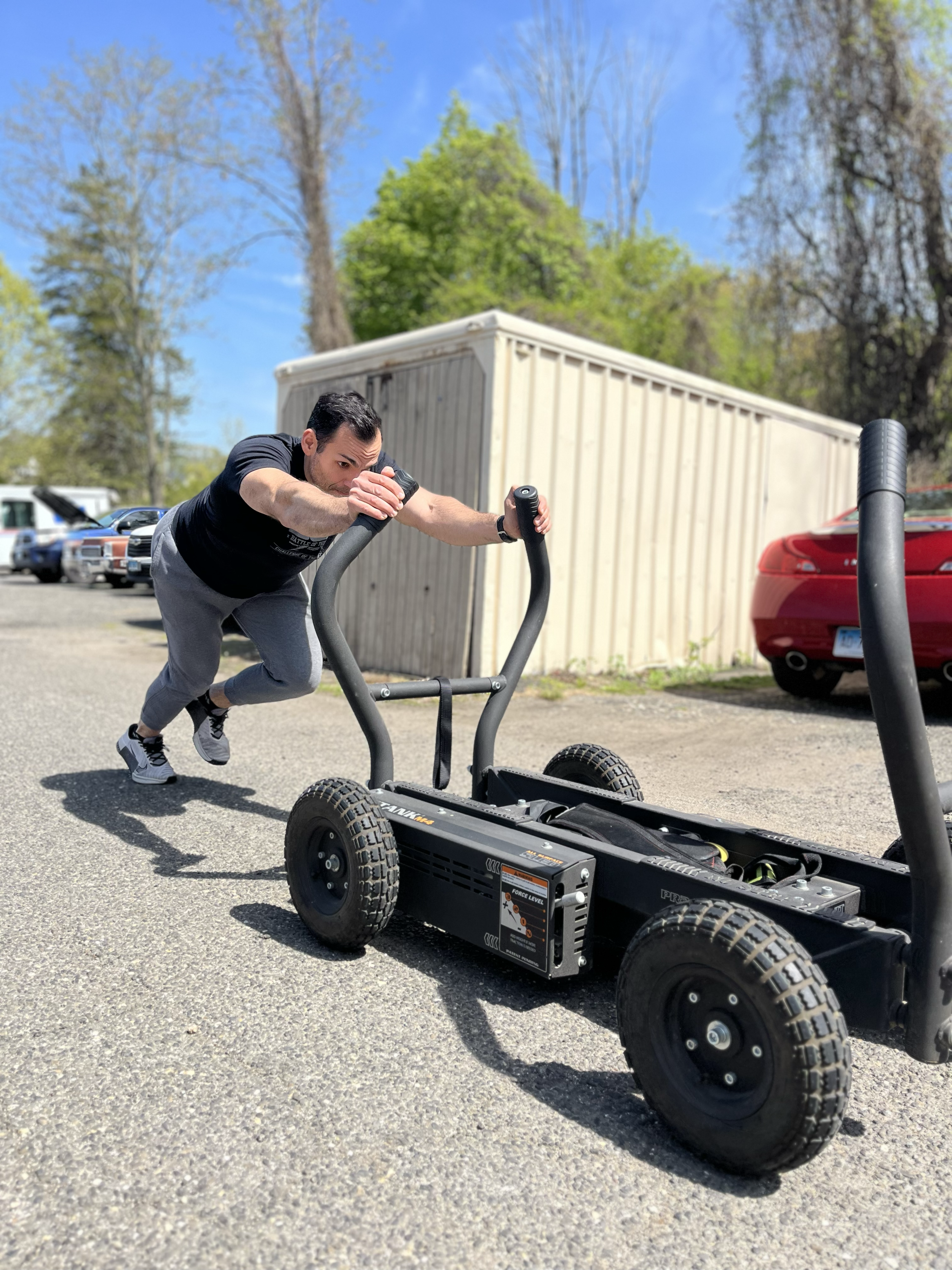 A man in a black shirt and gray pants pushes a black powered skateboard outdoors on a street with trees and several cars, including a red sports car and a beige container in the background.