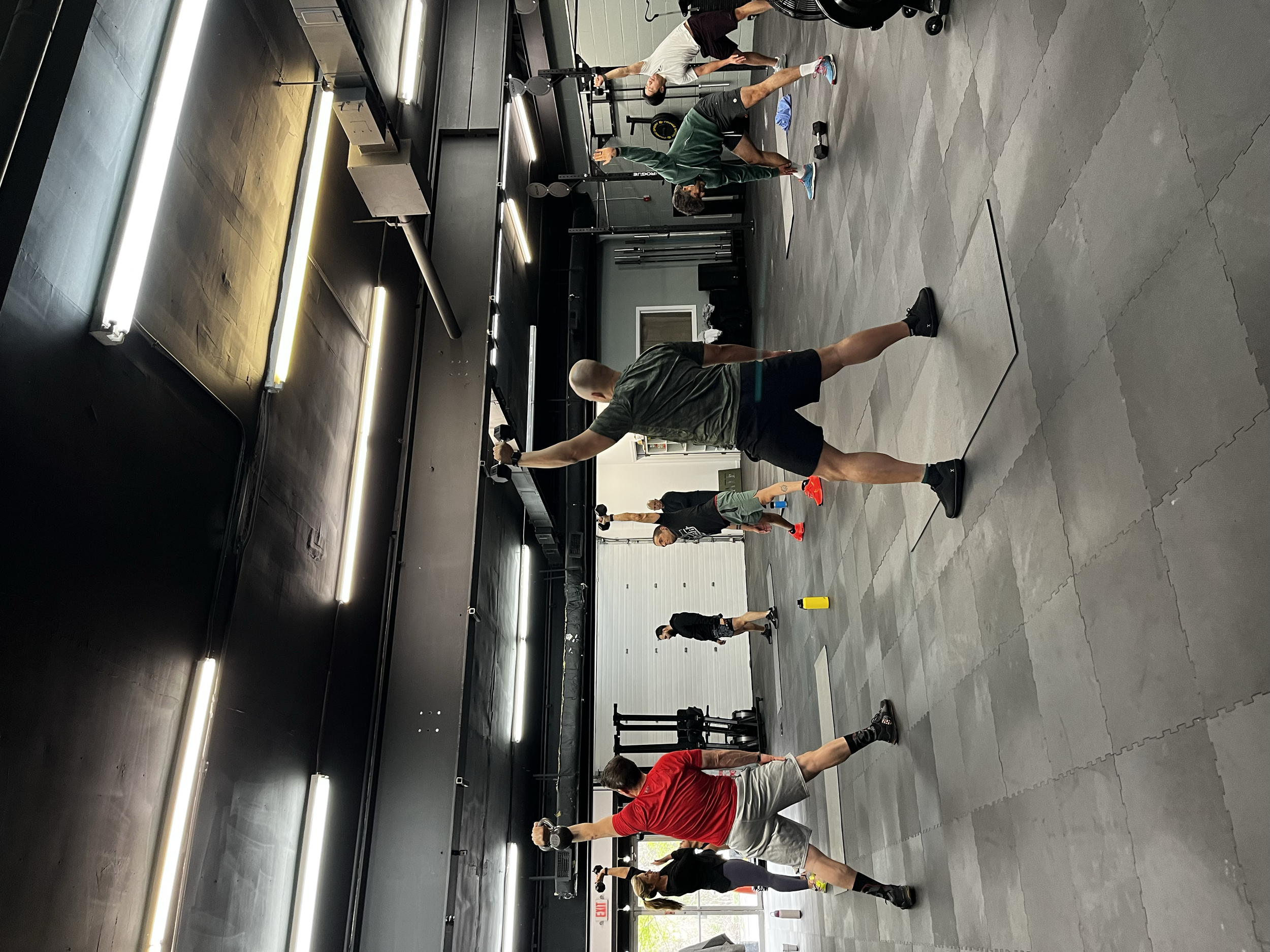 Several people in a gym are performing various exercises such as stretching and lifting weights on a black and gray gym floor. The workout area includes mirrors, gym equipment, and bright lighting.