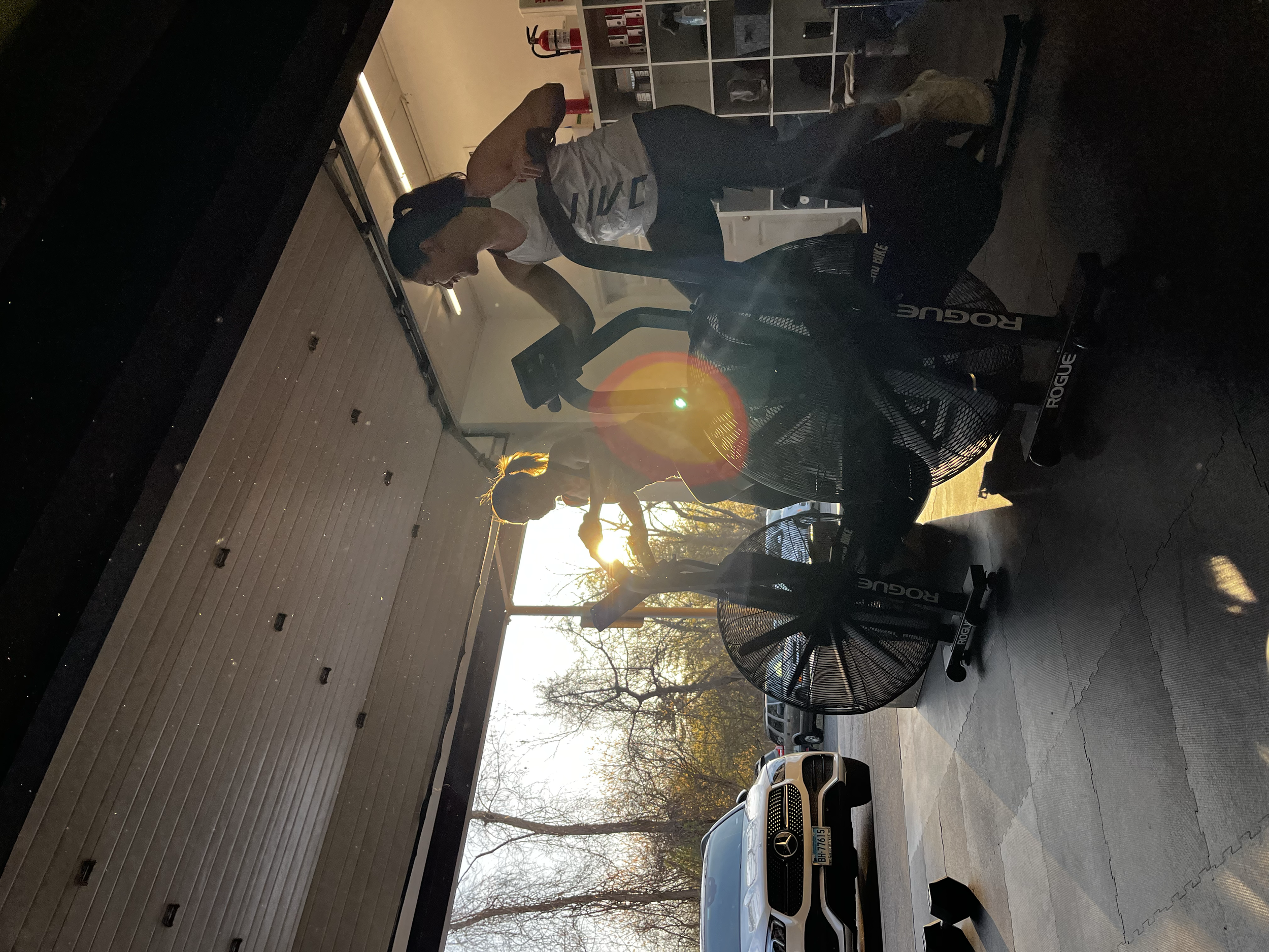 Inside a gym, a woman is exercising on a rowing machine while a man stands nearby. The sunlight is visible through large windows revealing garage doors, and trees outside. Various gym equipment is in the background.