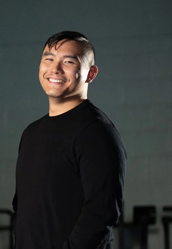 A smiling man with crossed arms, wearing a black CrossFit t-shirt, poses against a dark background with blue lighting.