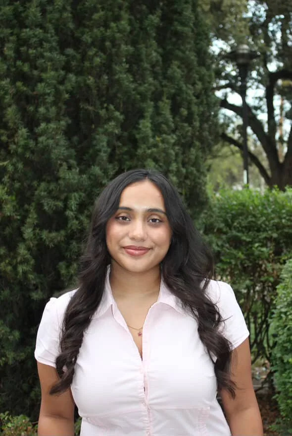 A woman with long dark hair and a necklace with a small star pendant, smiling indoors with a neutral background.
