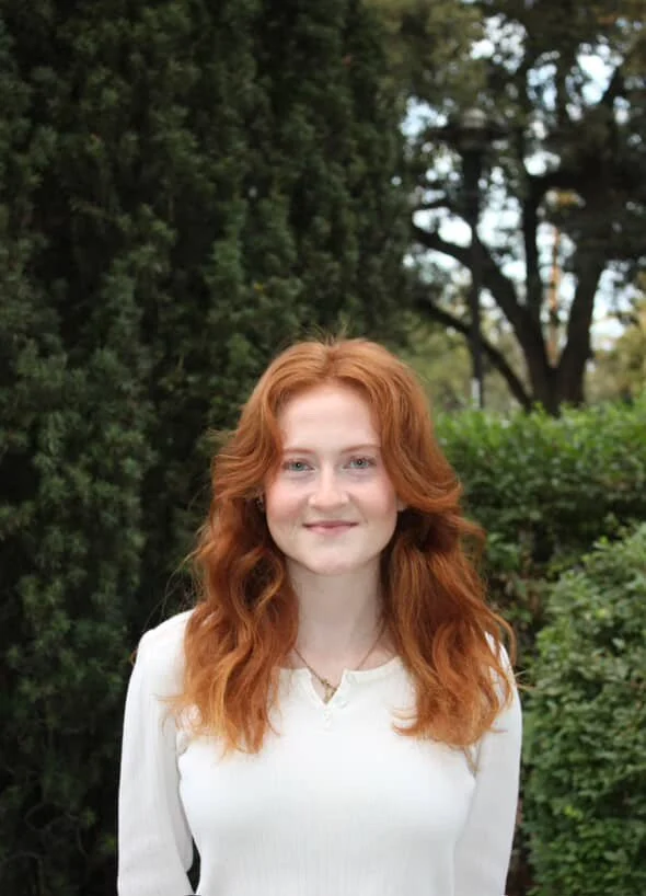 Woman with red hair in a floral dress standing in a sunny garden with palm trees in the background.