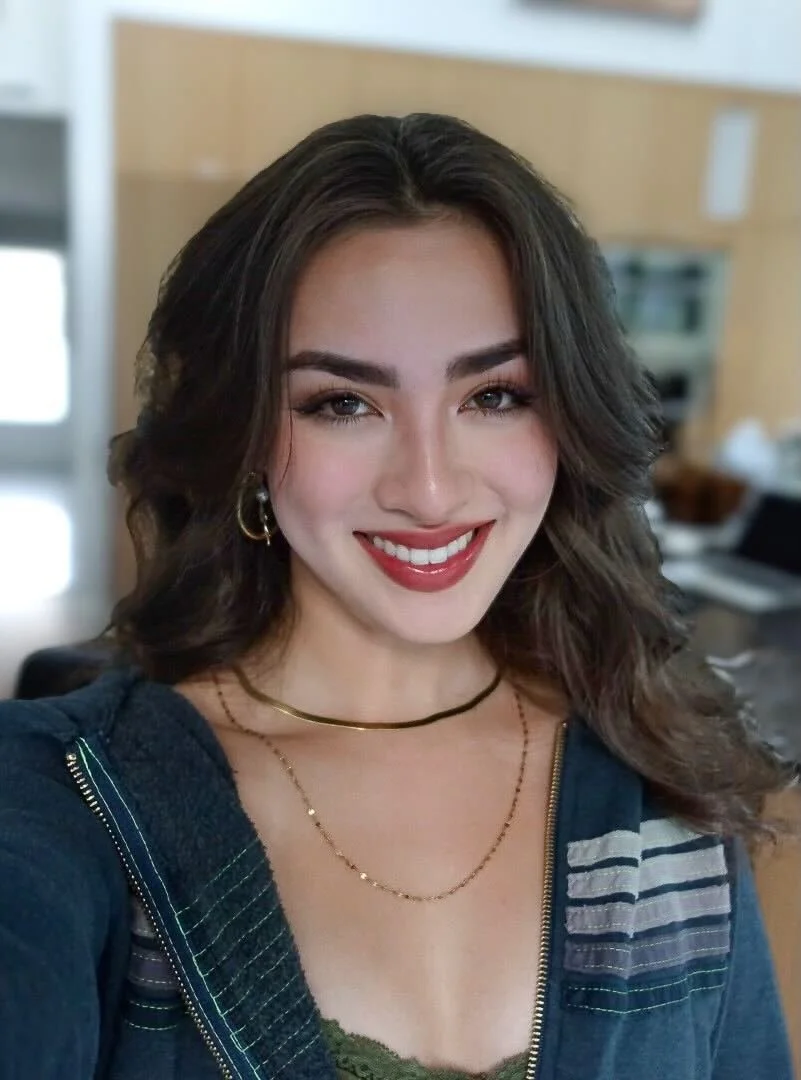 Smiling woman with wavy hair, wearing a dark jacket and layered necklaces, indoors.