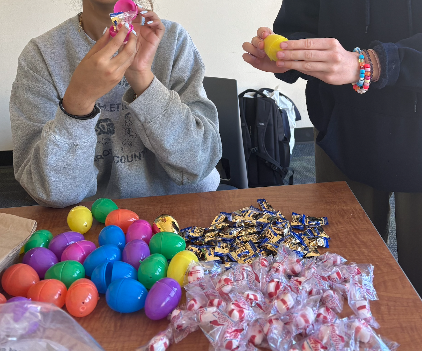 Two people filling colorful plastic eggs with candies on a wooden table. Various colored plastic eggs and wrapped candies are scattered on the table.