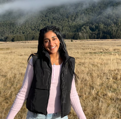 Person in outdoor setting with dry grass, forest background, wearing black vest and pink long-sleeve shirt, smiling.