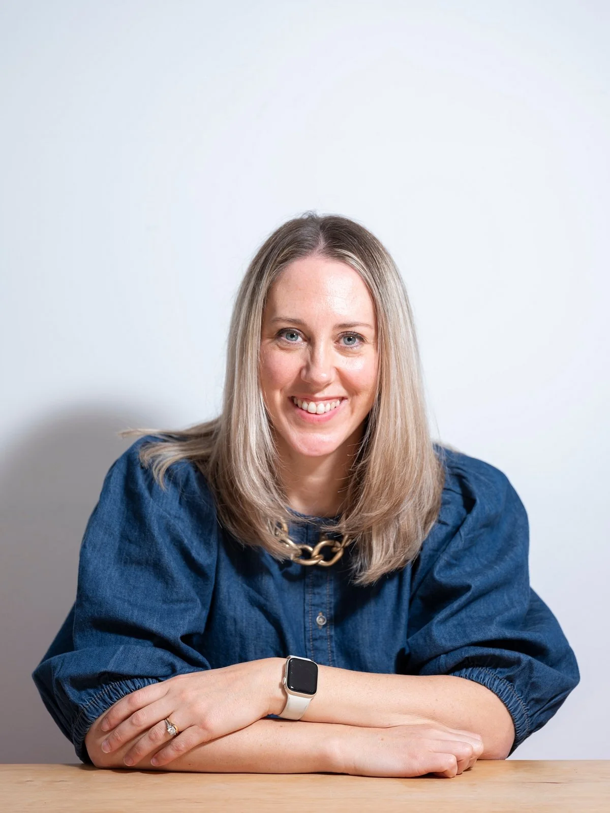 A woman with blonde hair smiling and sitting at a wooden table, wearing a blue denim shirt, a chunky necklace, and a smartwatch.