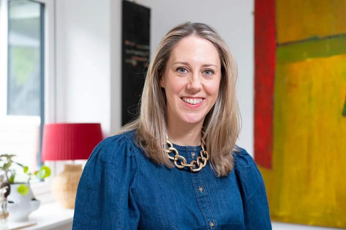 A smiling woman with shoulder-length blonde hair, wearing a blue denim top and a chunky gold necklace, standing inside a room with a window, red lampshade, and colorful abstract painting on the wall.