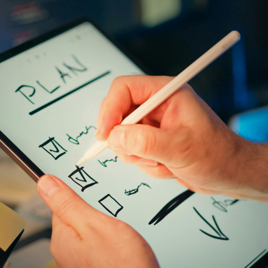 A hand writing the word Plan on a notepad surrounded by sketched ideas
