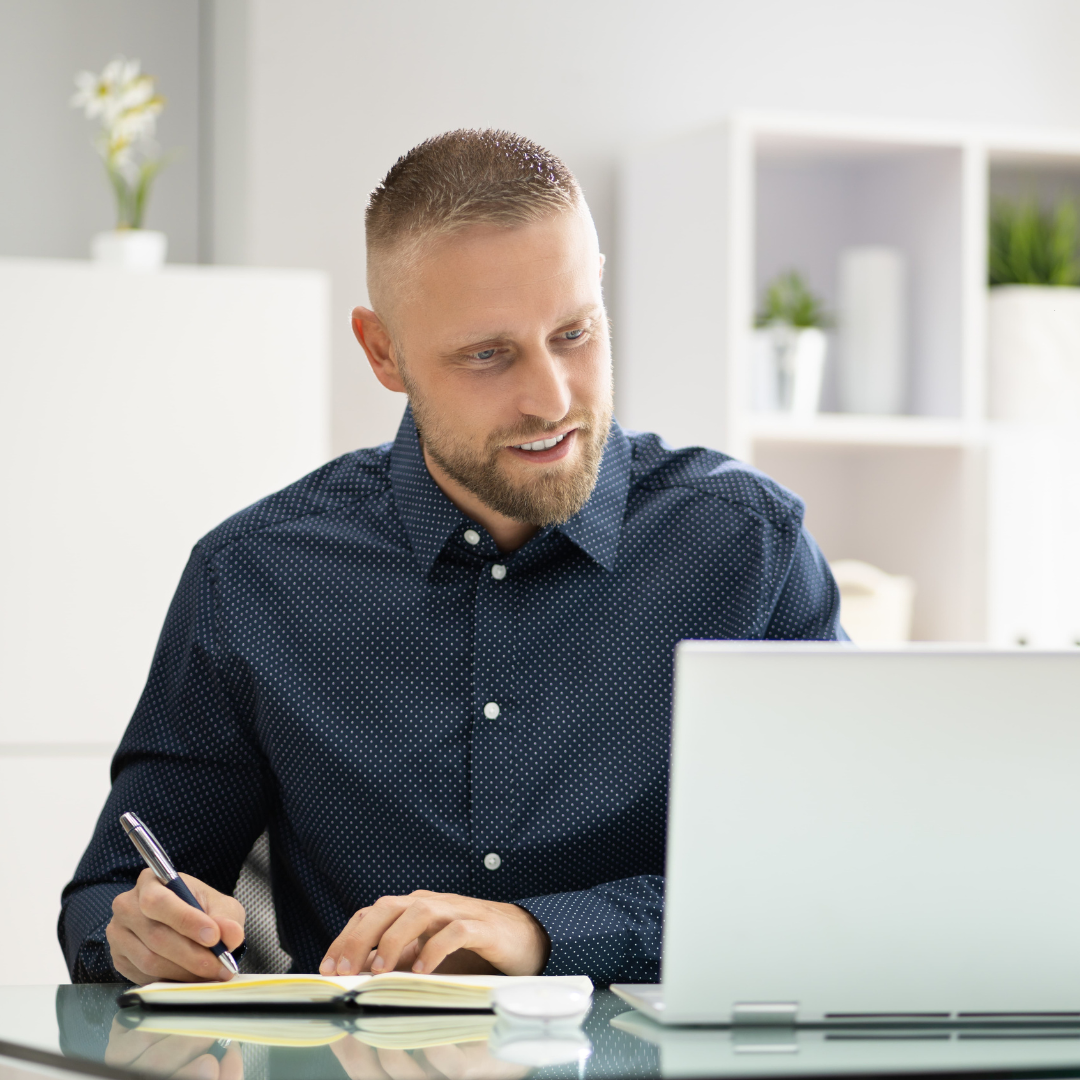 A man taking notes beside his laptop at a bright desk