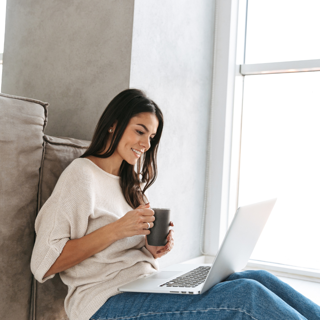 A woman smiling at her laptop with a cup of coffee, sitting by a sunlit window