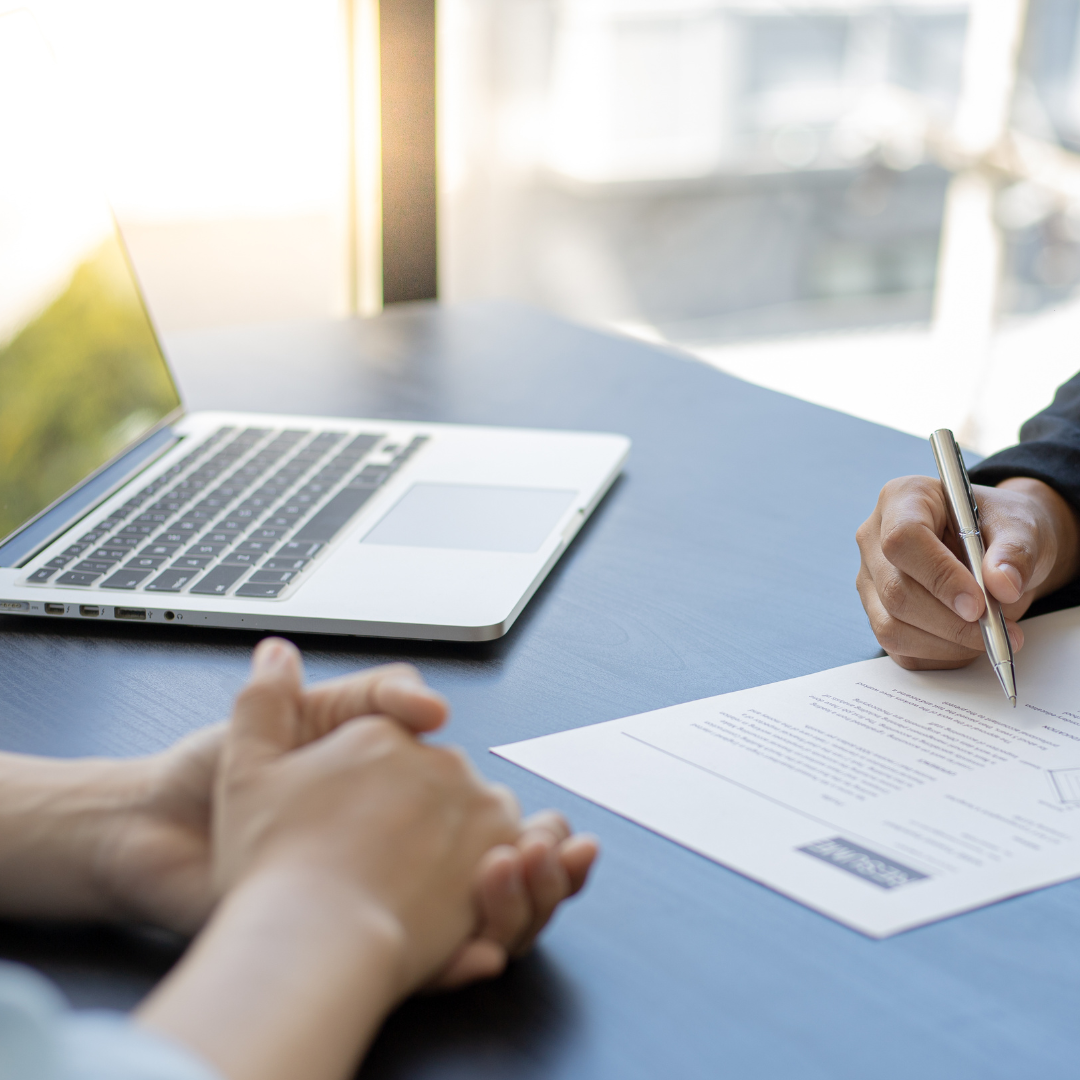 Two people meeting at a desk, one signing a document beside an open laptop