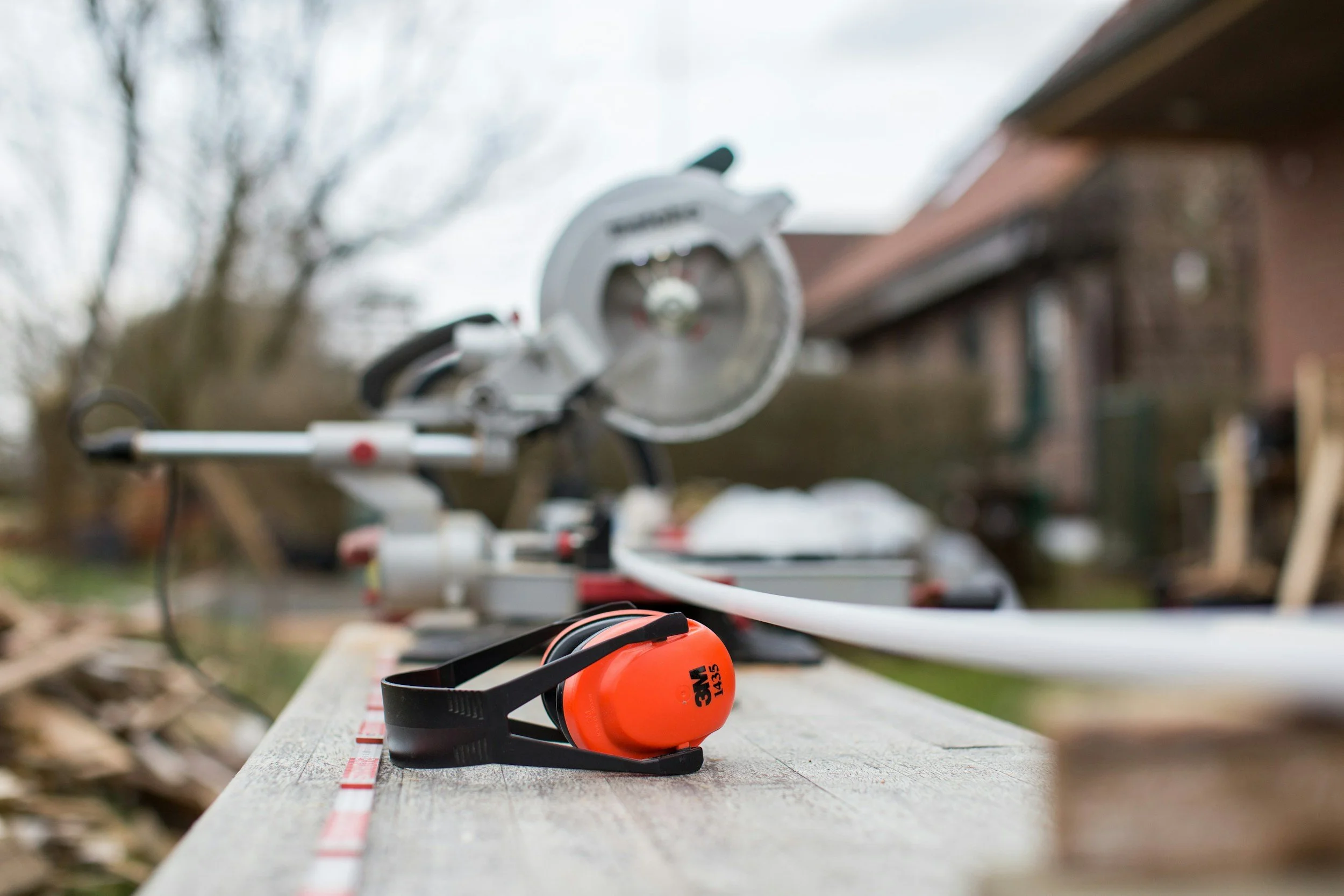 A construction worker's tool setup on a wooden workbench outdoors, including a circular saw and orange earmuffs, with a blurred background of a house and trees.