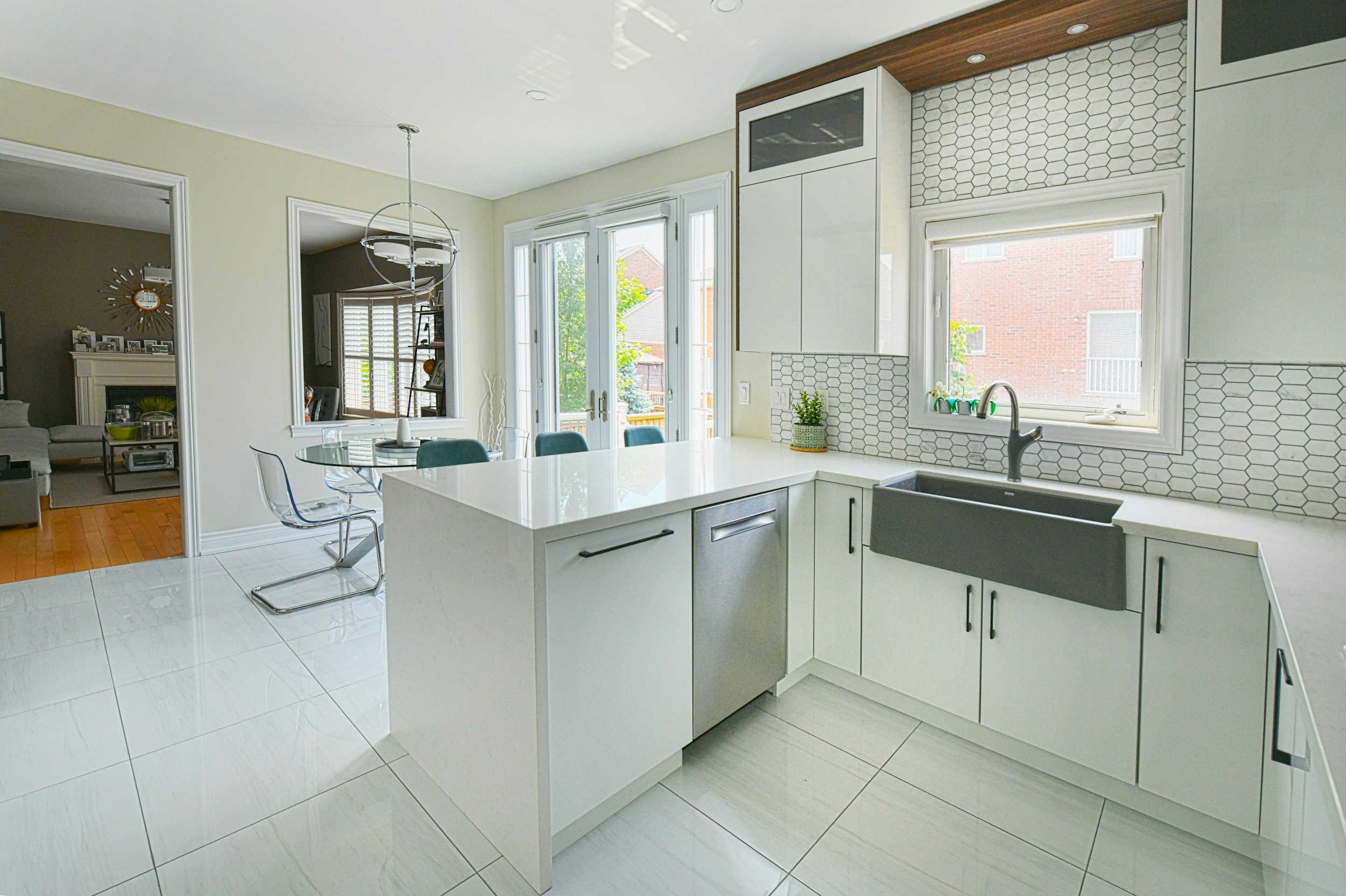 Modern kitchen with white cabinets, a gray farmhouse sink, white tile backsplash, and a small potted plant on the counter. Large window above the sink, glass doors leading outside, and a dining area with glass table and chairs.