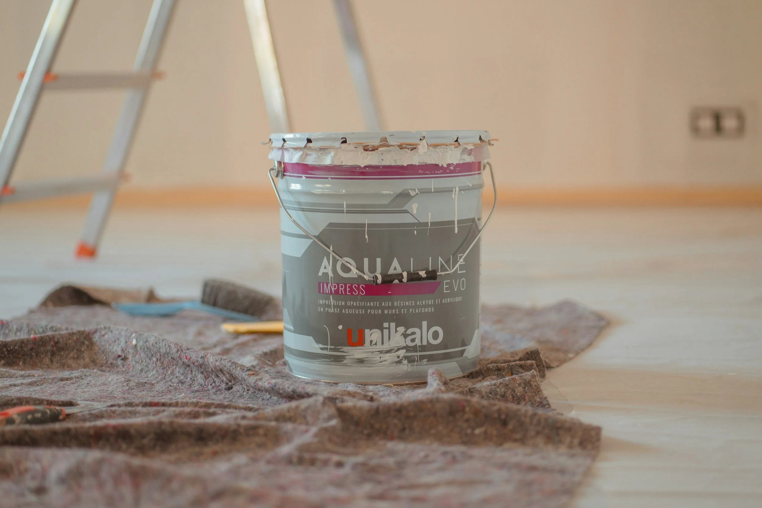 A paint bucket with grey and pink paint, placed on a plastic sheet in a room under renovation, with a ladder in the background.