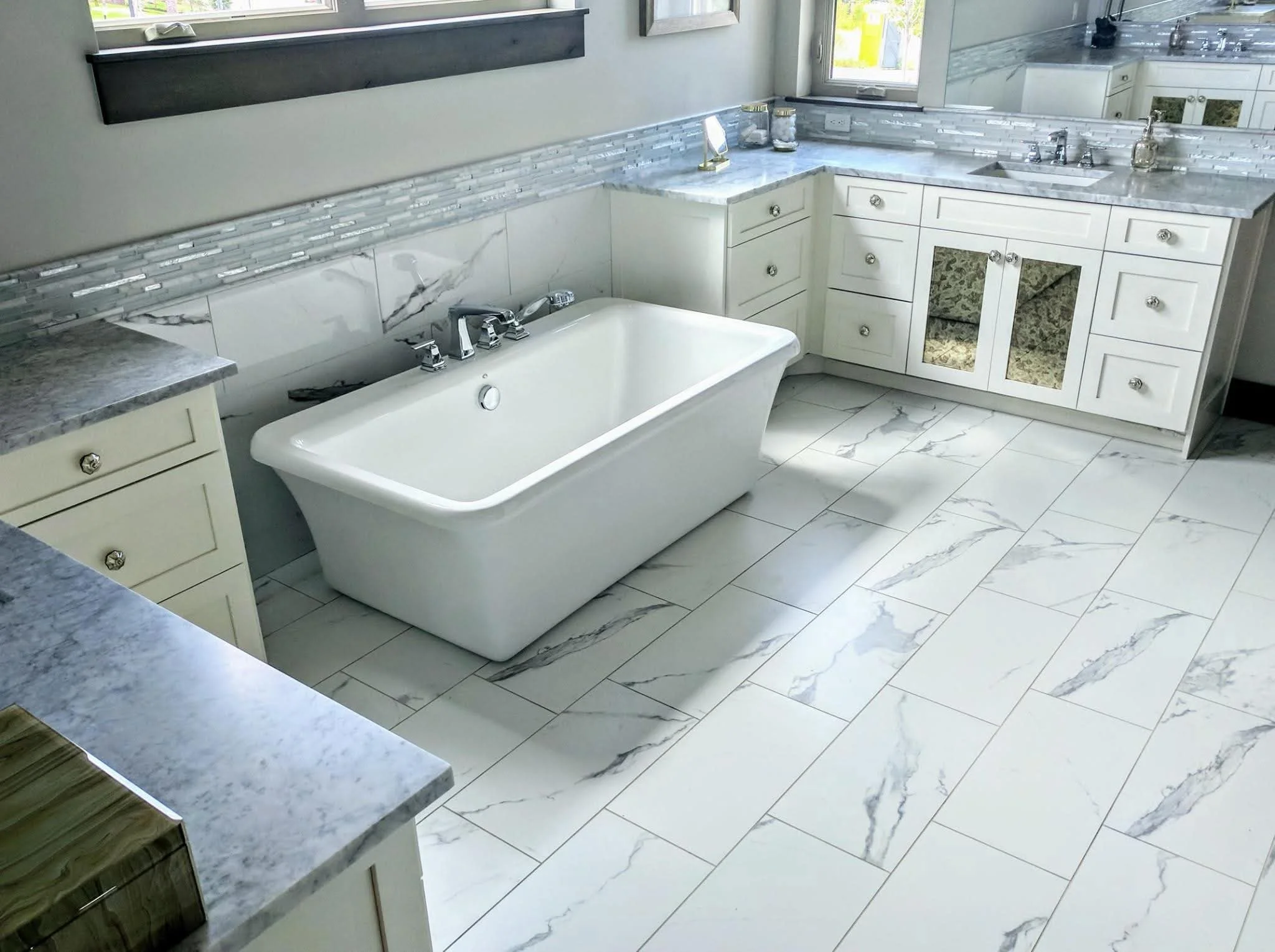 A modern bathroom with a white free-standing bathtub, marble-like tile floors, and white cabinetry with glass-paneled doors. There is a window above the bathtub and a marble countertop with a sink and various toiletries.