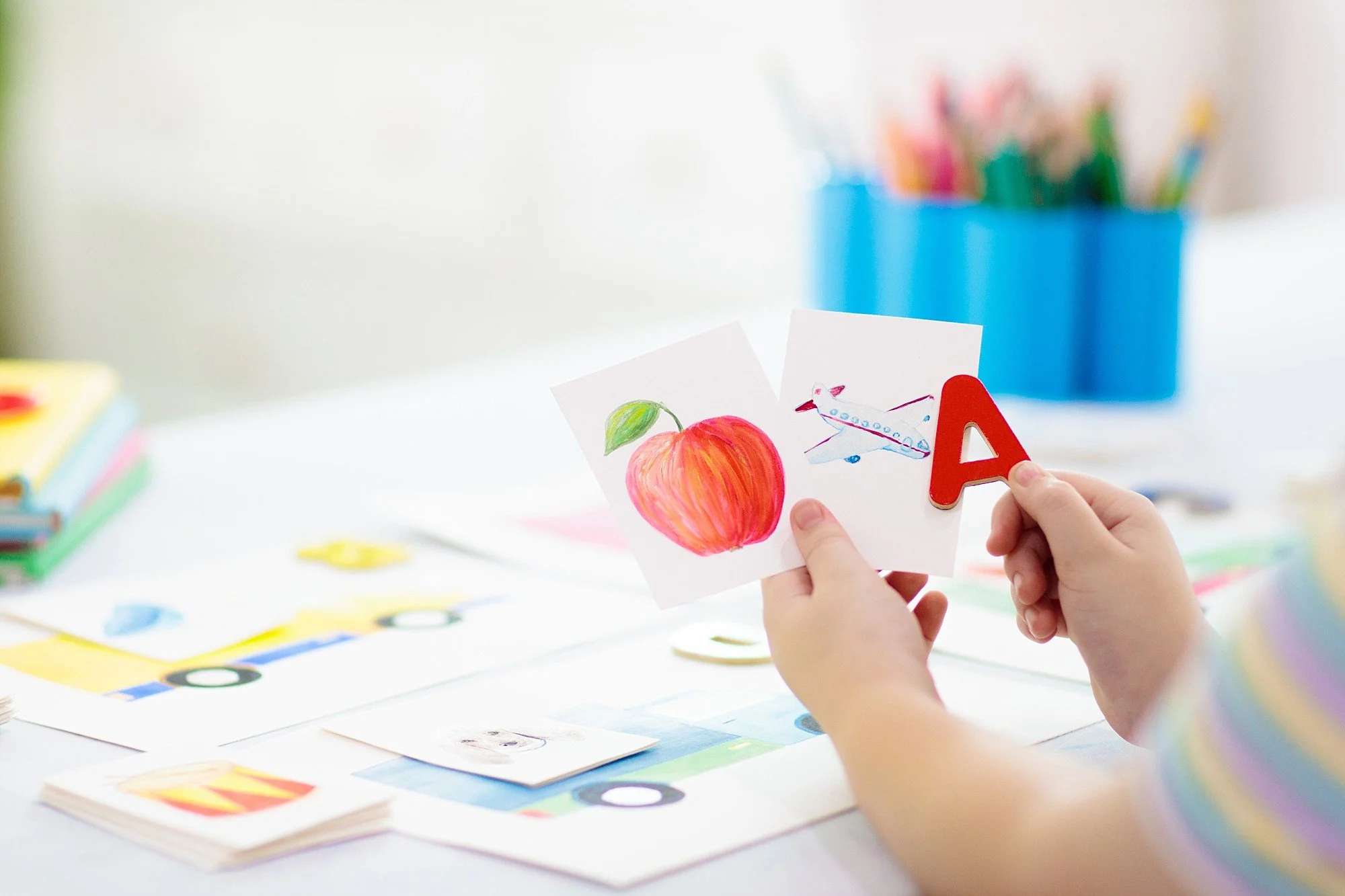 Child's hands holding flashcards with a drawing of an apple and an airplane, with a red letter 'A' held in front of the cards, on a table with arts and crafts supplies.