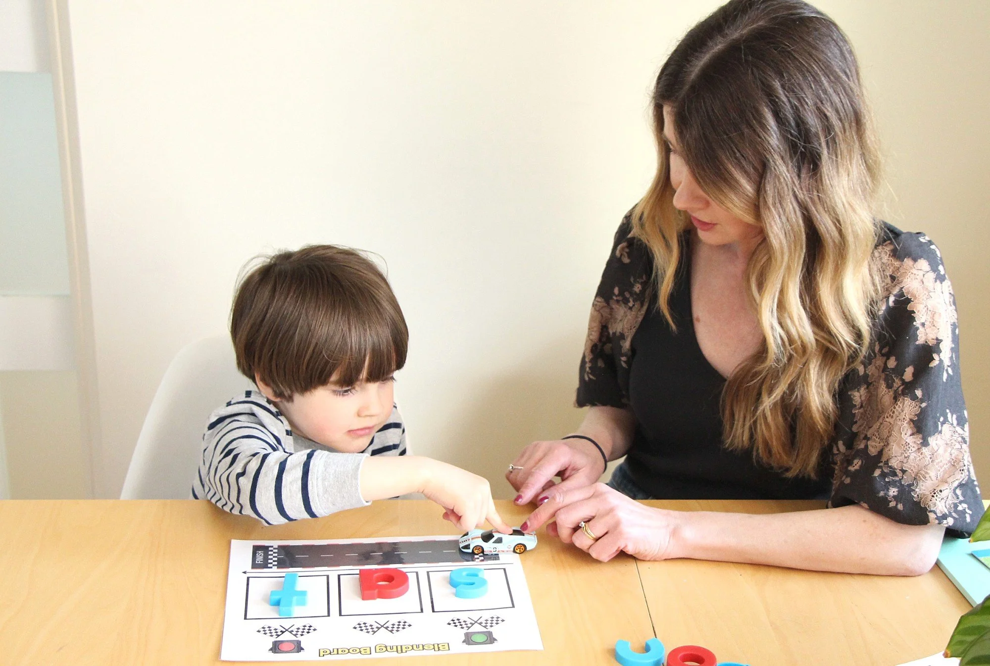 A young boy and a woman playing a road safety game with toy cars and foam letter blocks at a wooden table.