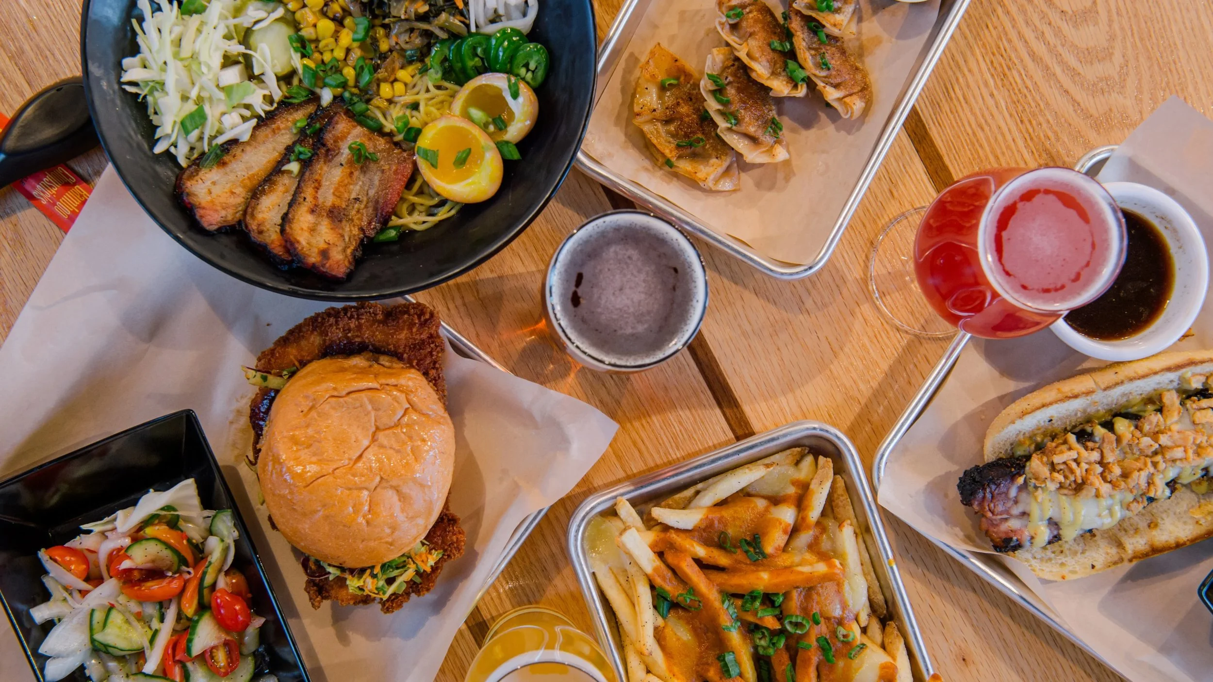 A table of various Asian and American dishes, including a bowl of ramen with grilled pork and boiled eggs, two types of gyoza, a cheeseburger with fried chicken, a slice of sandwich with fried chicken, fries, and cheese, salads, and multiple glasses 