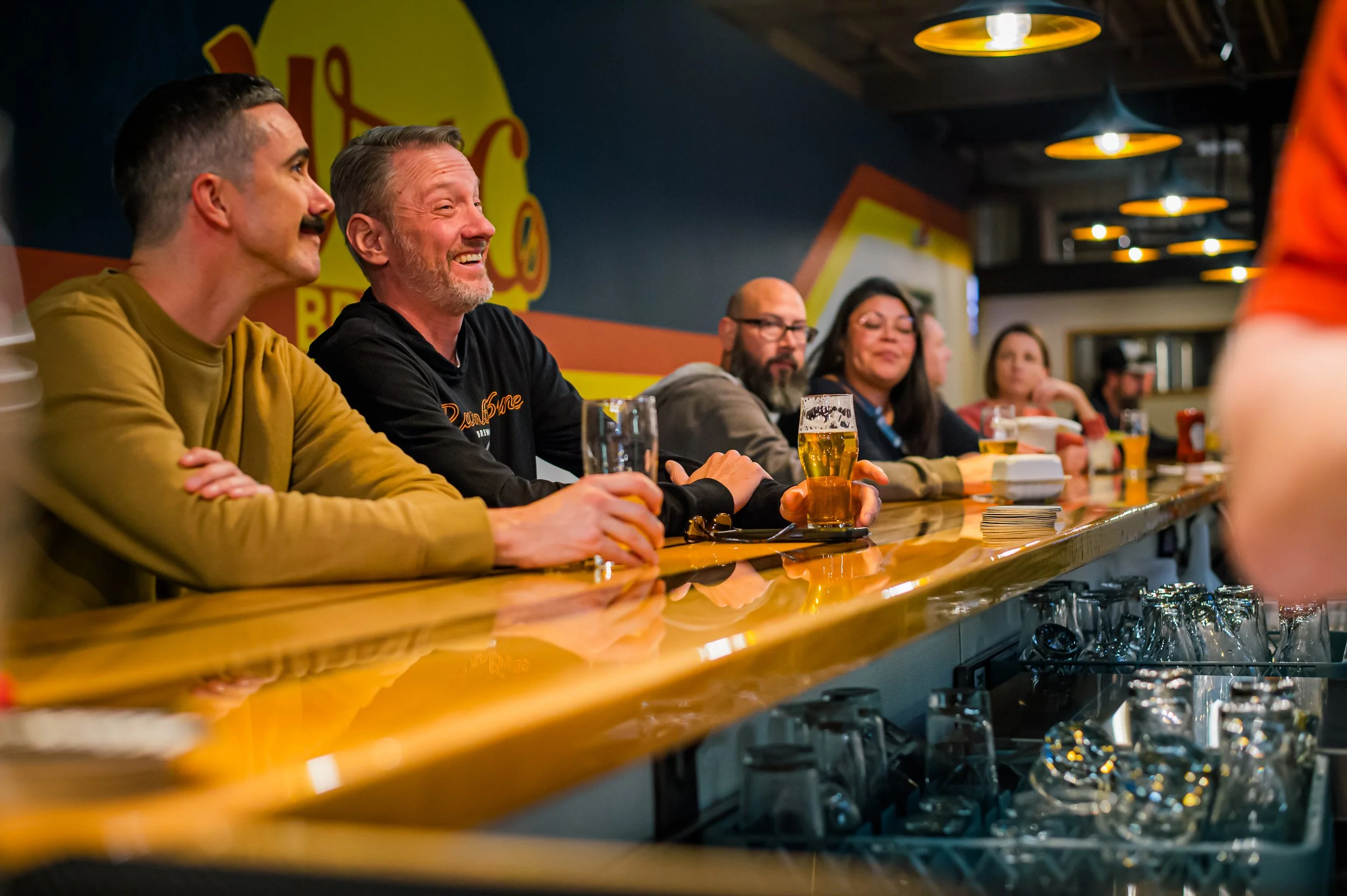 A group of people sitting at a bar with drinks, engaging and smiling, in a lively pub or bar setting.