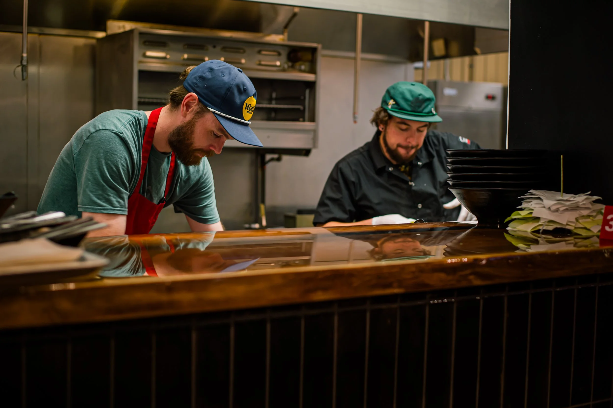 Two men working behind a wooden counter in a restaurant kitchen, both wearing hats and focused on their tasks.