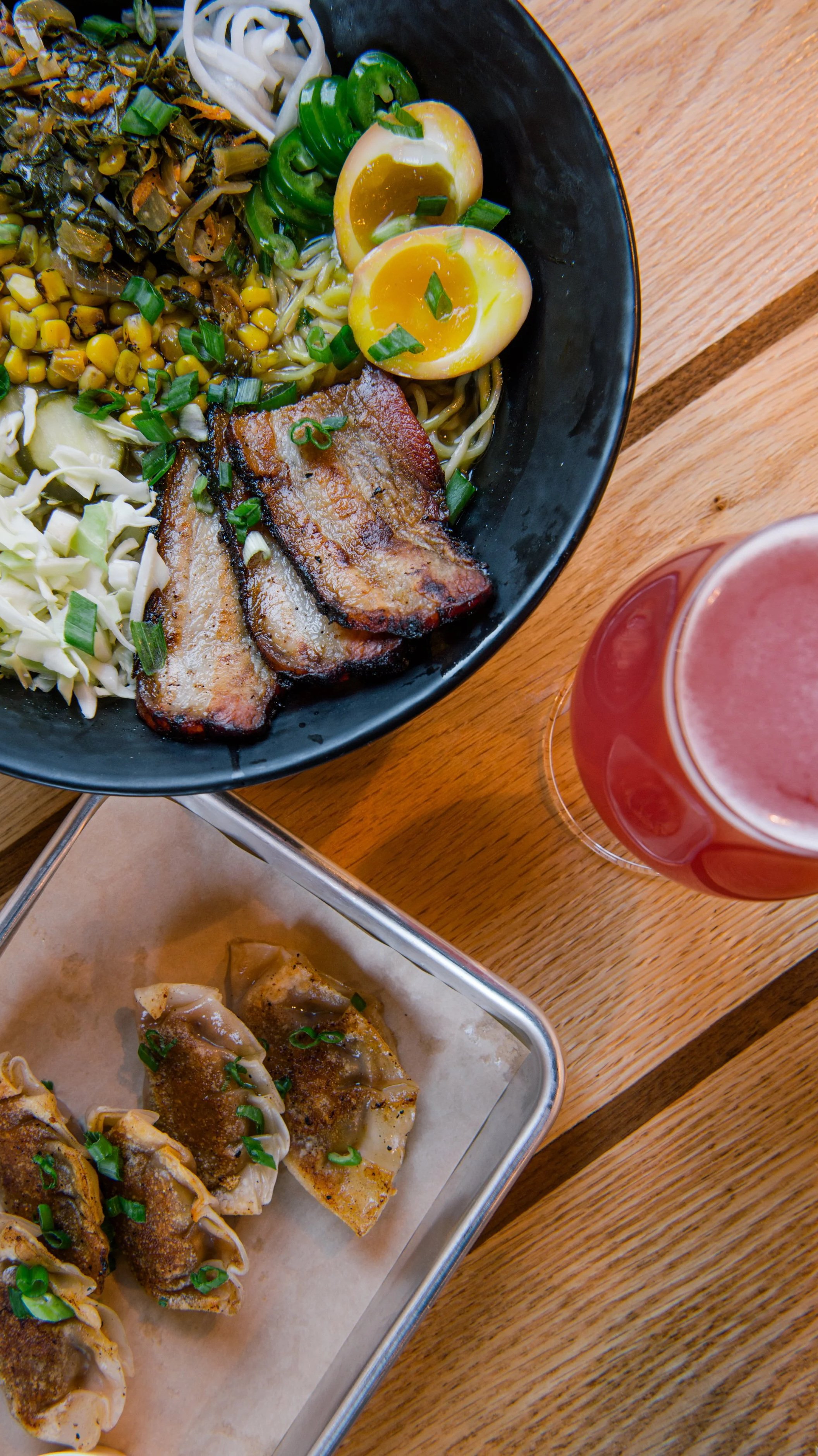 A bowl of ramen with sliced pork, soft boiled eggs, green onions, corn, seaweed, and noodles, with a pink drink nearby. A small tray of fried dumplings with green onions on top is also visible.