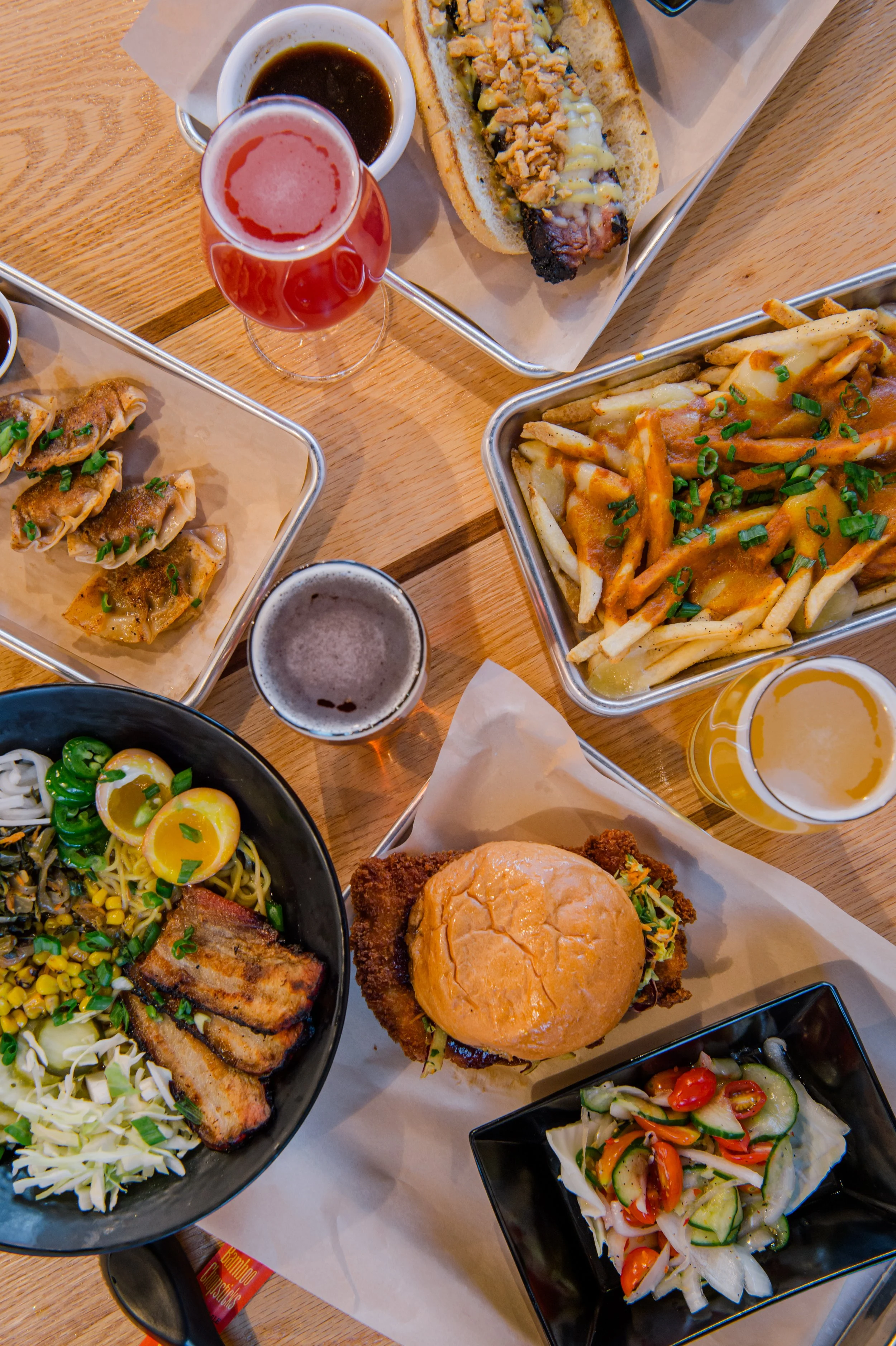 A table with various dishes including a hot dog with toppings, a bowl of fries topped with cheese and green onions, a burger with fried chicken, a bowl of Asian noodles with sliced eggs and pork, a small salad with cucumber and cherry tomatoes, and several cups of different colored beers.
