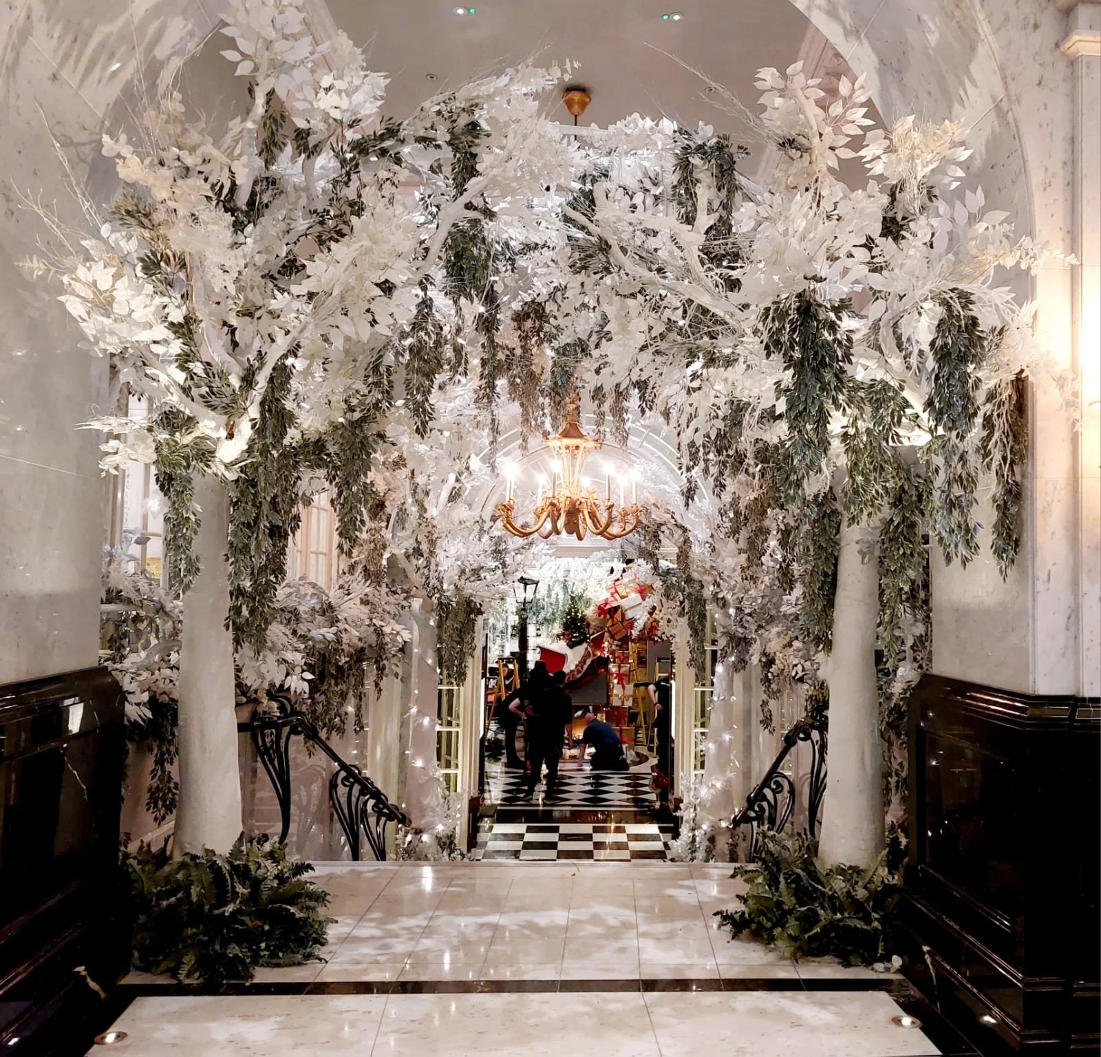 Interior decorated with white trees, green foliage, and string lights leading to a Christmas scene with a Christmas tree and presents, lit by a chandelier and wall lamps.