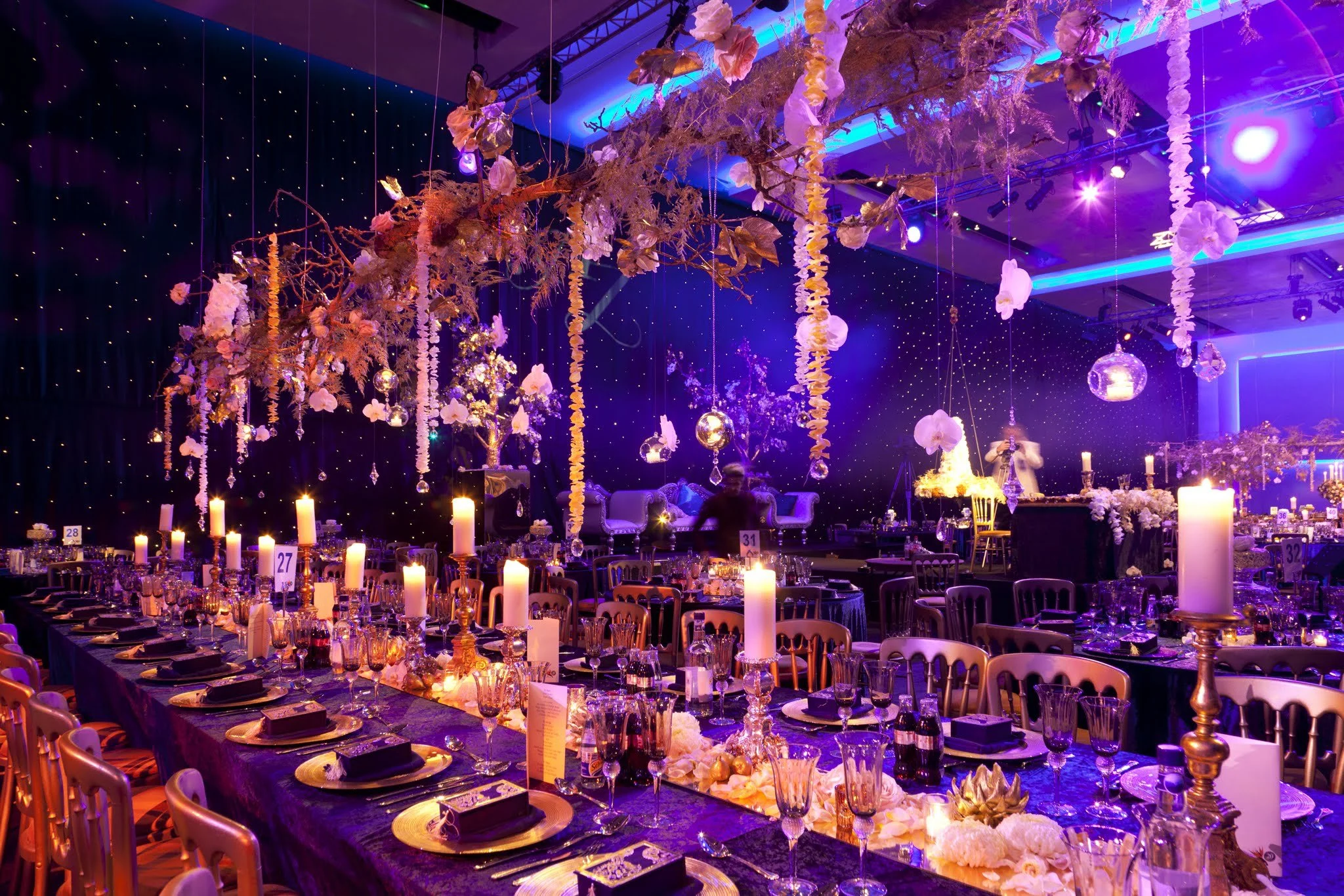 Elegant banquet hall decorated with purple lighting, floral arrangements, hanging white flowers, candles, and ornate table settings for a celebration or wedding reception.