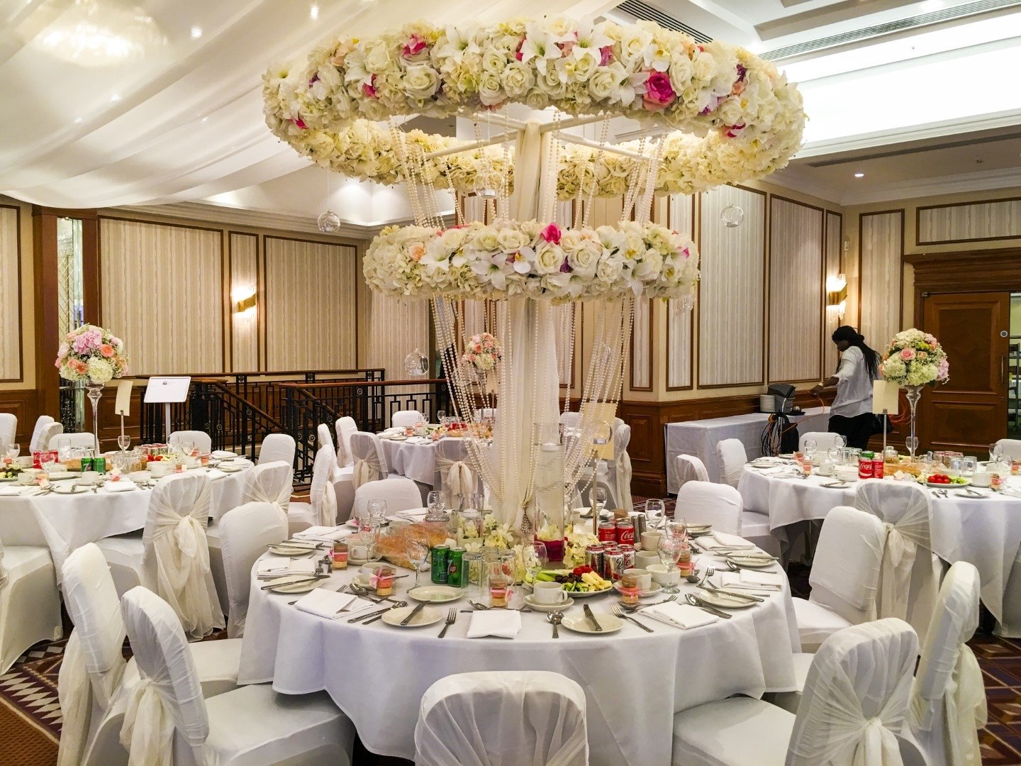 Elegant banquet hall with round tables decorated with white tablecloths and floral centerpieces, featuring a large floral chandelier made of white and pink flowers hanging from the ceiling.