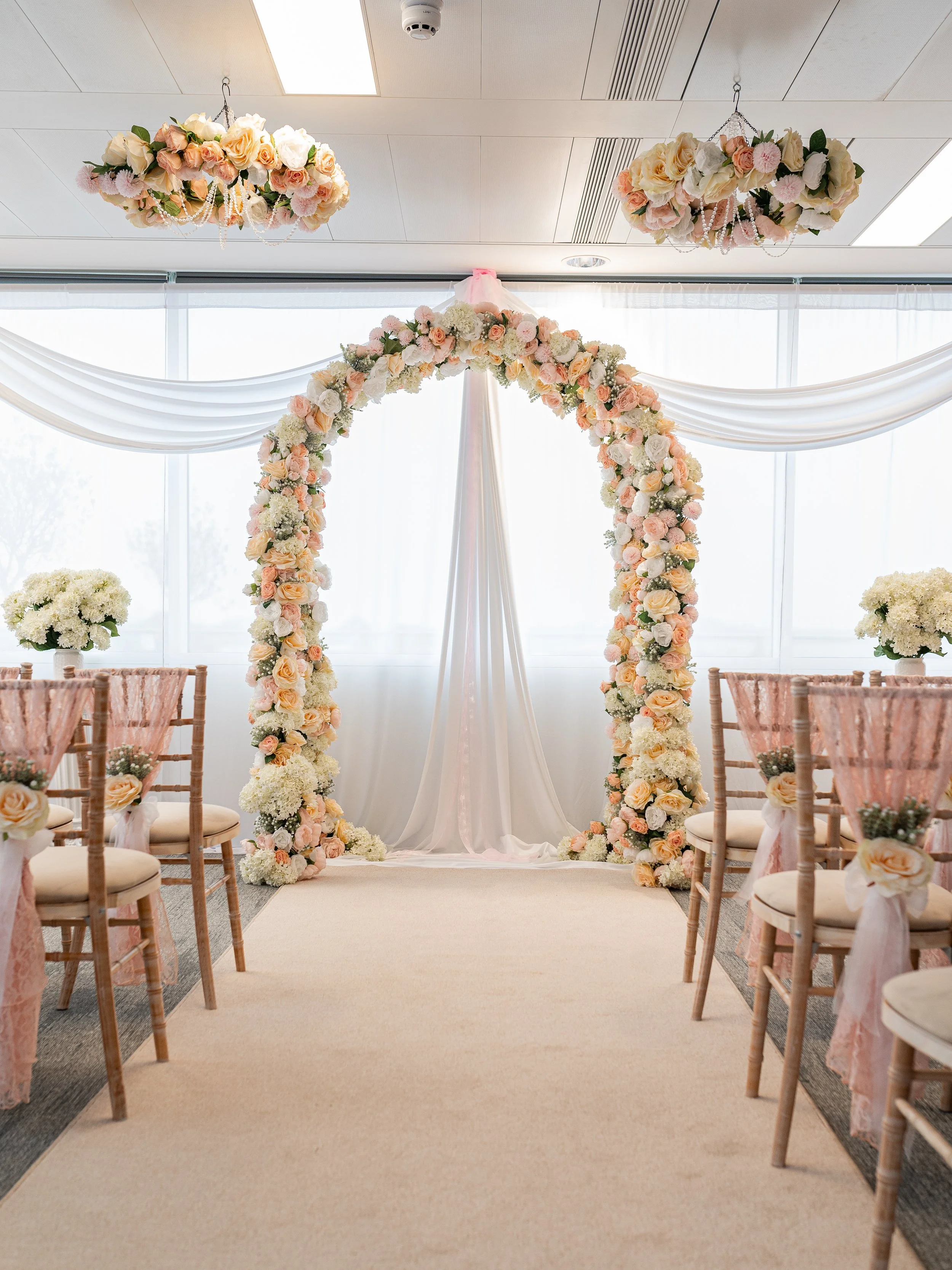 A wedding ceremony arch decorated with pink, white, and peach flowers, with sheer pink fabric draped behind it, surrounded by chairs with pink sashes and floral accents, in a bright room with large windows.