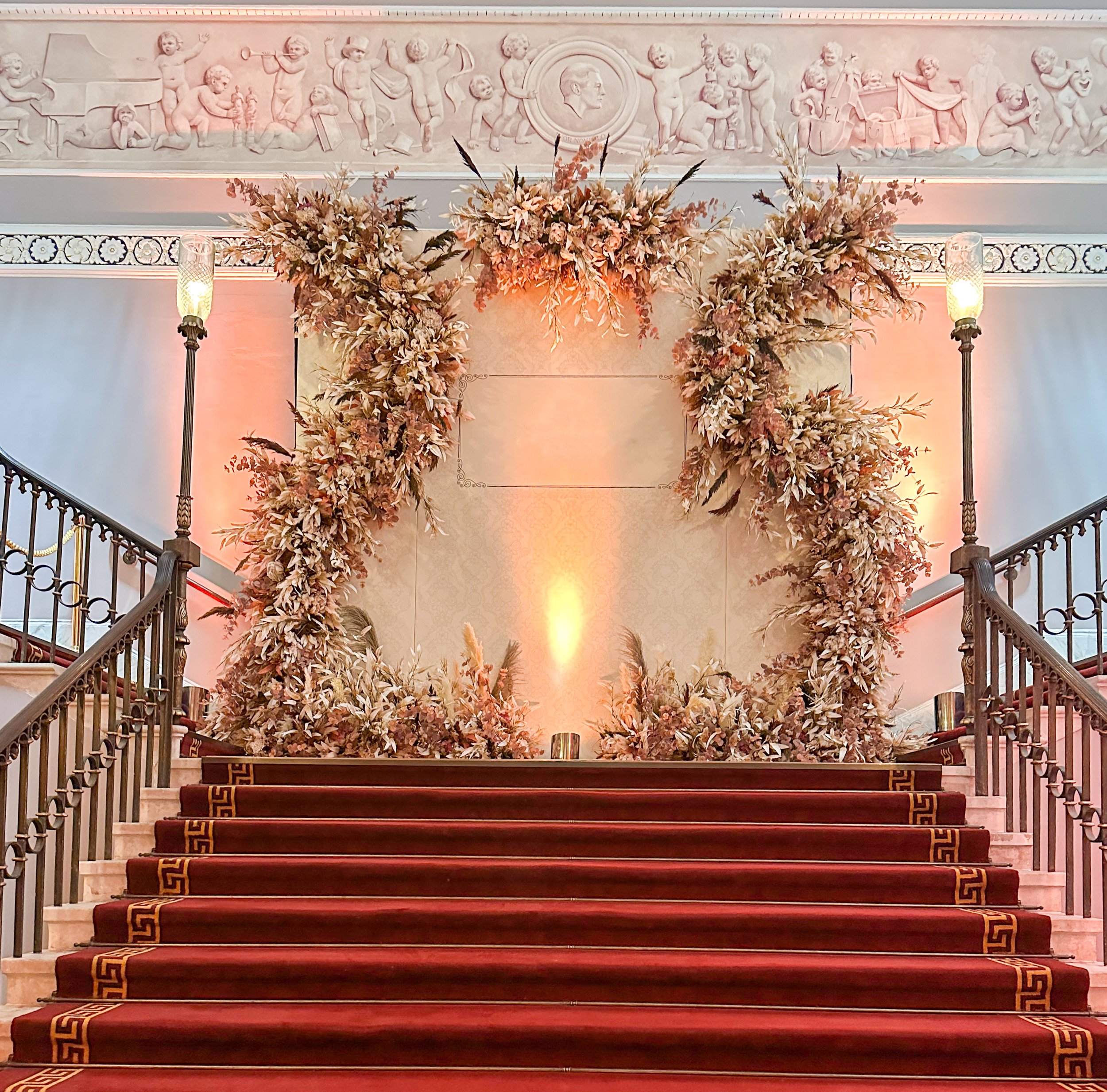 Elegant staircase with red carpet leading up to an ornate floral arch of dried pink and beige flowers, with decorative wall sculptures and soft lighting.