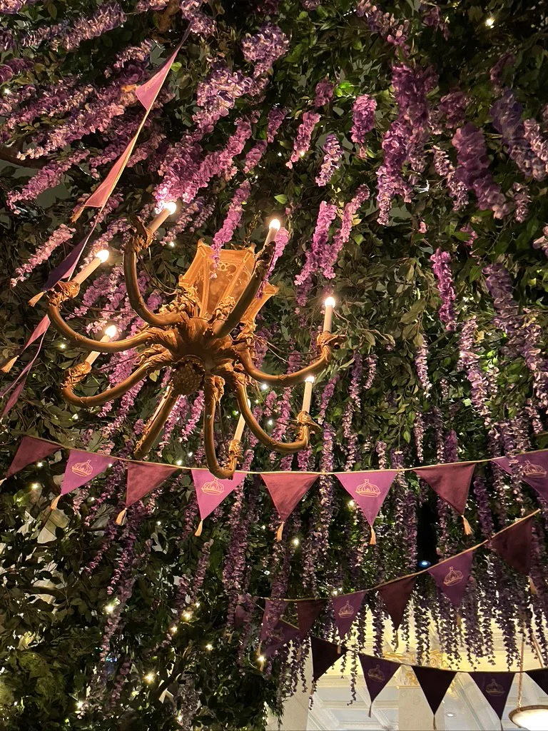 A decorated ceiling with purple hanging flowers, a gold chandelier with lit candles, and pink triangular banners with cupcake icons.