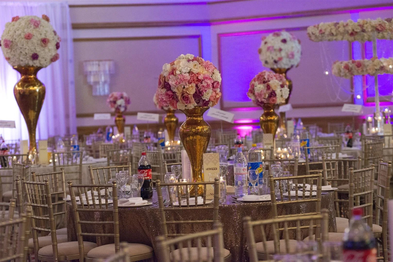 Elegant event seating with gold chairs, decorated with pink and white floral centerpieces on tall gold vases, set in a banquet hall with purple lighting.