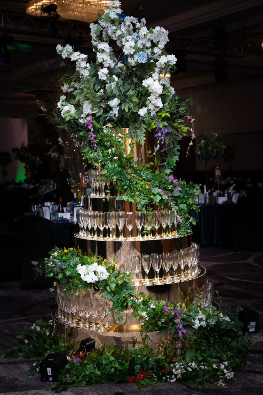 A tall floral centerpiece with white, purple, and pink flowers, green foliage, and champagne glasses arranged on a multi-tiered gold stand, set in a banquet hall for a celebration or wedding.