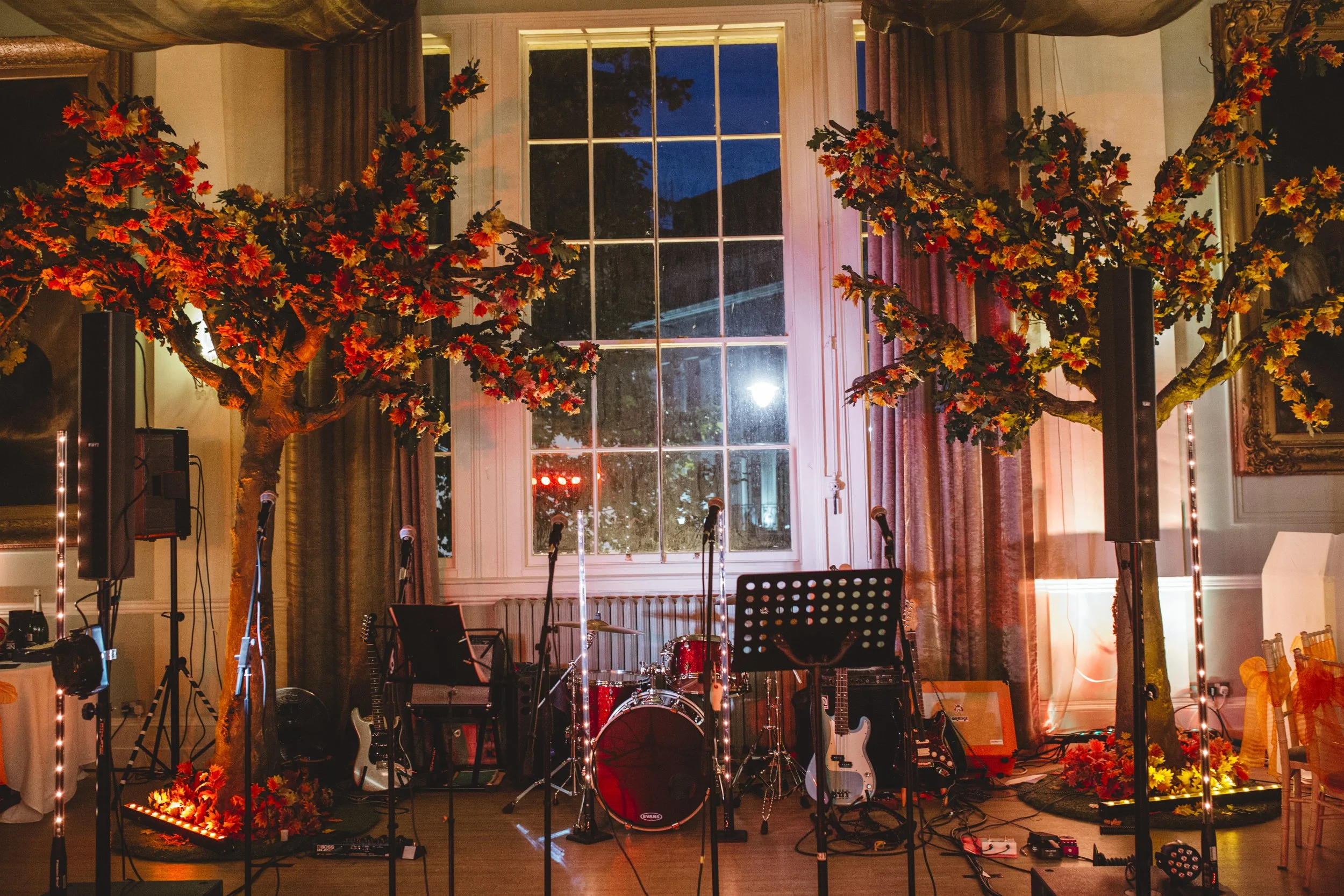 Indoor event setting with a stage featuring musical instruments, two artificial trees with autumn-colored leaves, and decorative lighting, in front of a large window showing an evening sky.