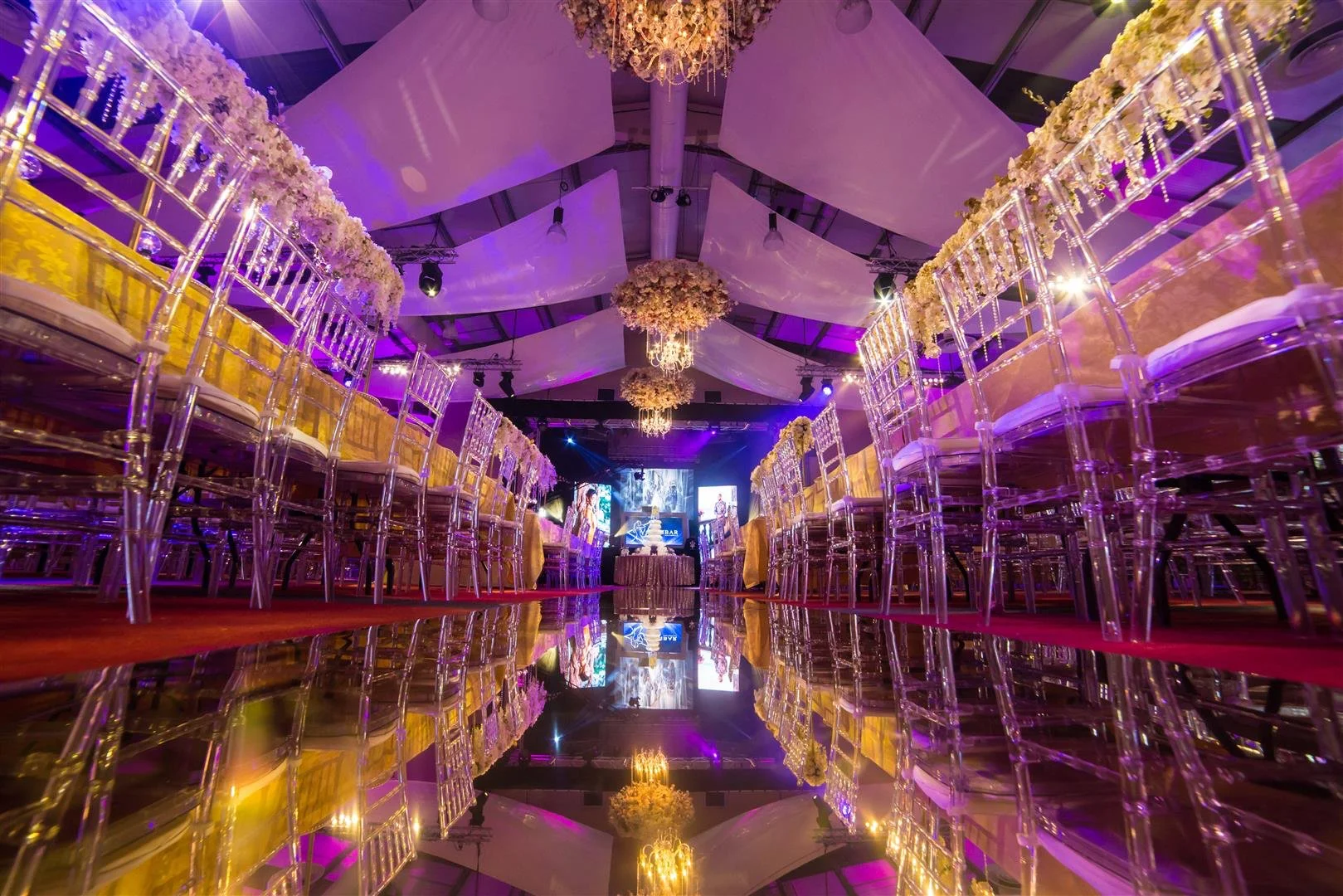 Elegant event hall decorated with purple and yellow lighting, floral arrangements, and transparent chairs with yellow cushions, set for a formal celebration or wedding reception.