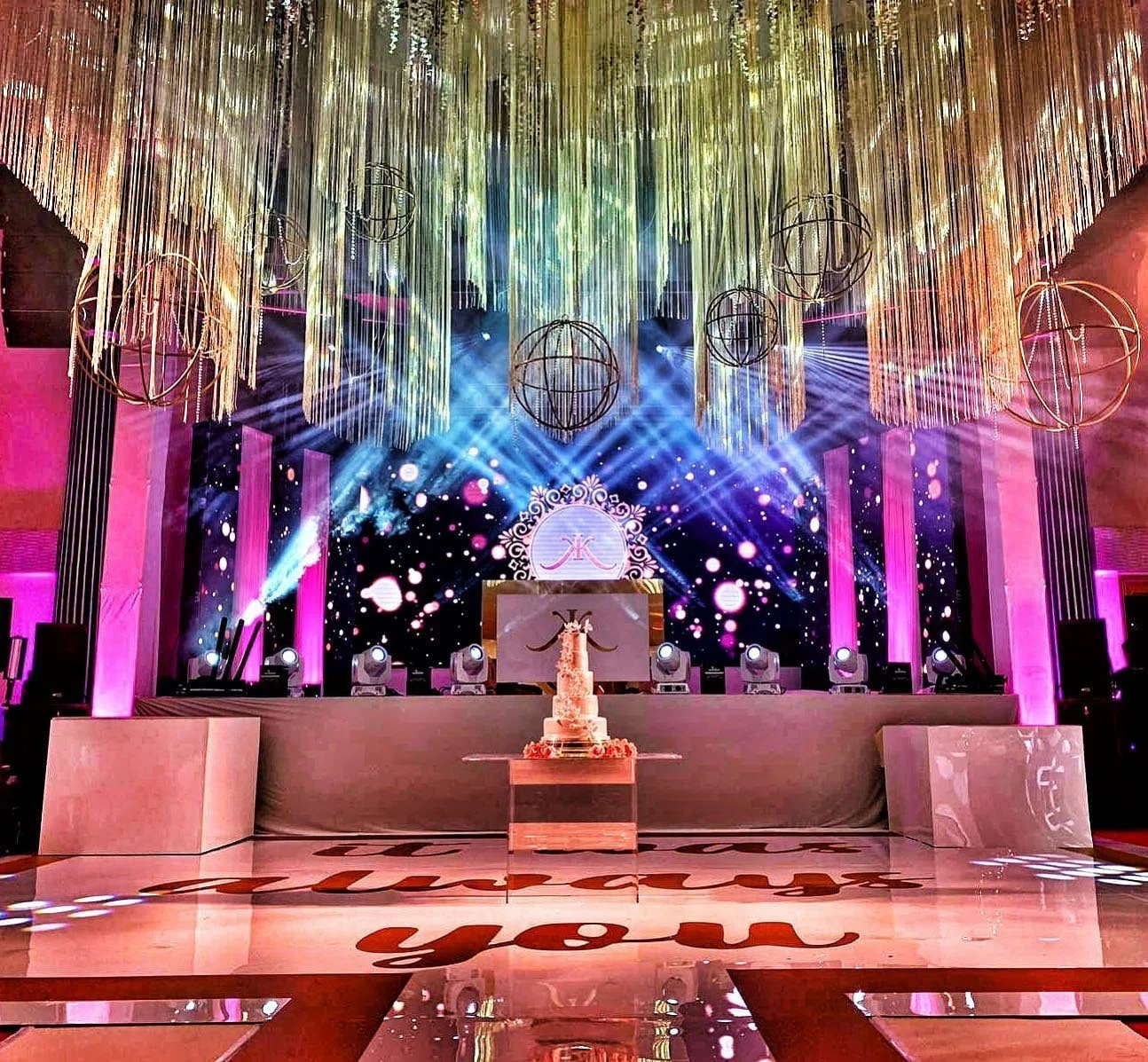 Elegant event stage with a tiered wedding cake in front, surrounded by pink lighting, hanging decorations, and a large digital screen displaying bright lights and graphics.