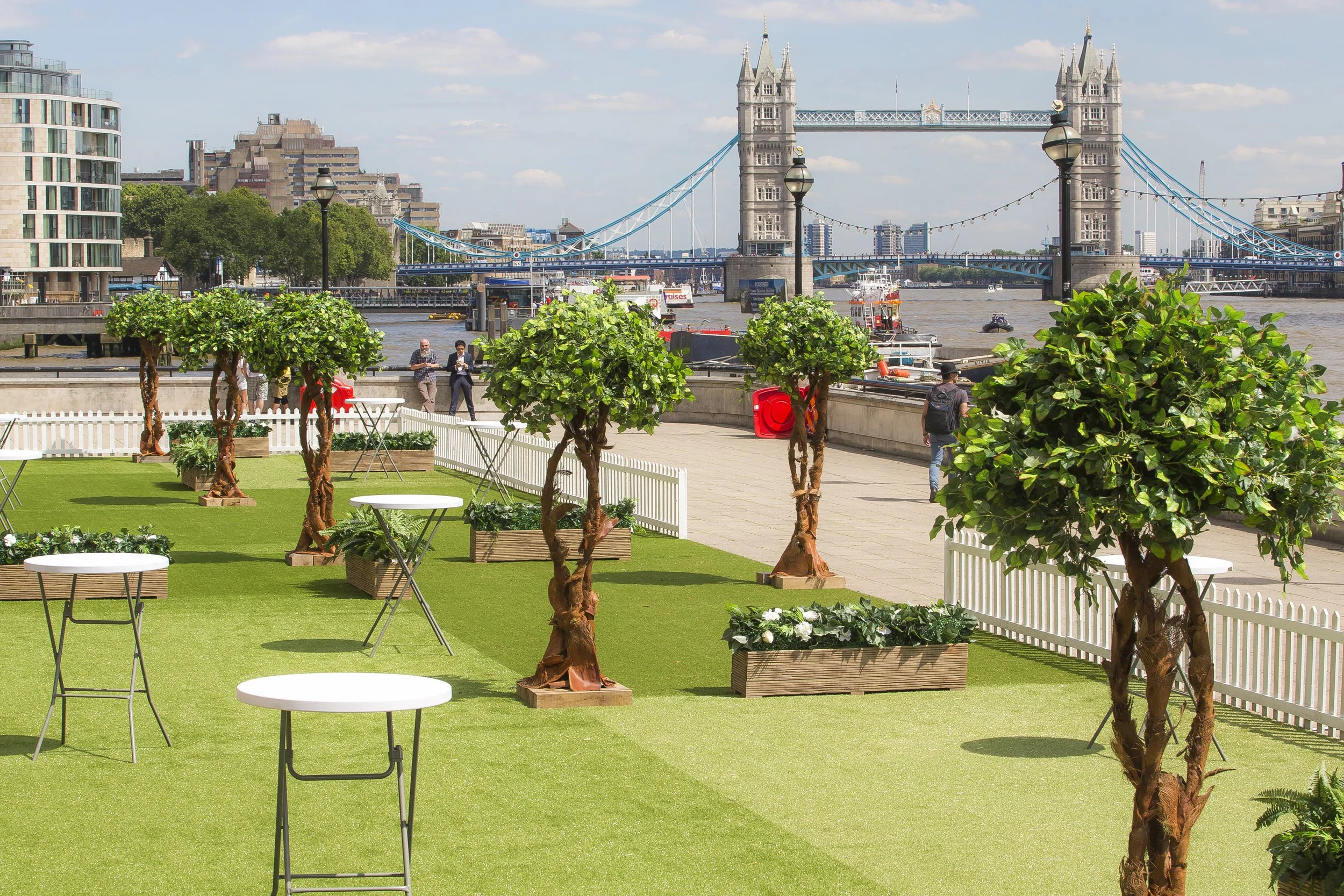 Outdoor patio with potted trees, tables, and a view of Tower Bridge over the River Thames in London.