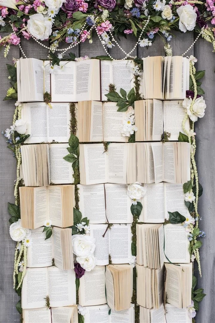Open books arranged vertically surrounded by green leaves, white and purple flowers, and decorative beads on a gray wooden background.